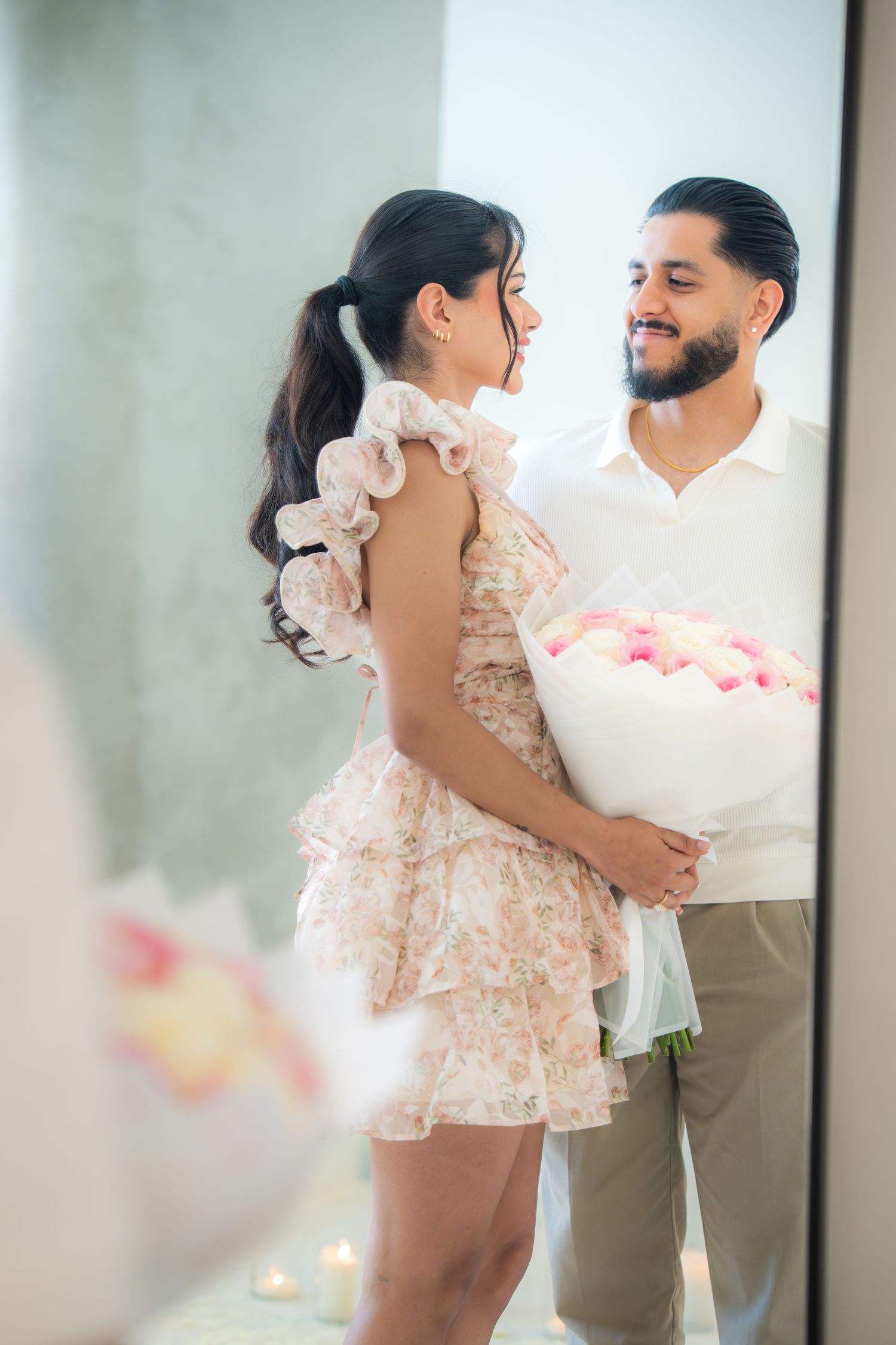 Couple sharing a romantic moment, man holding a bouquet of pink and white roses in a softly lit room.