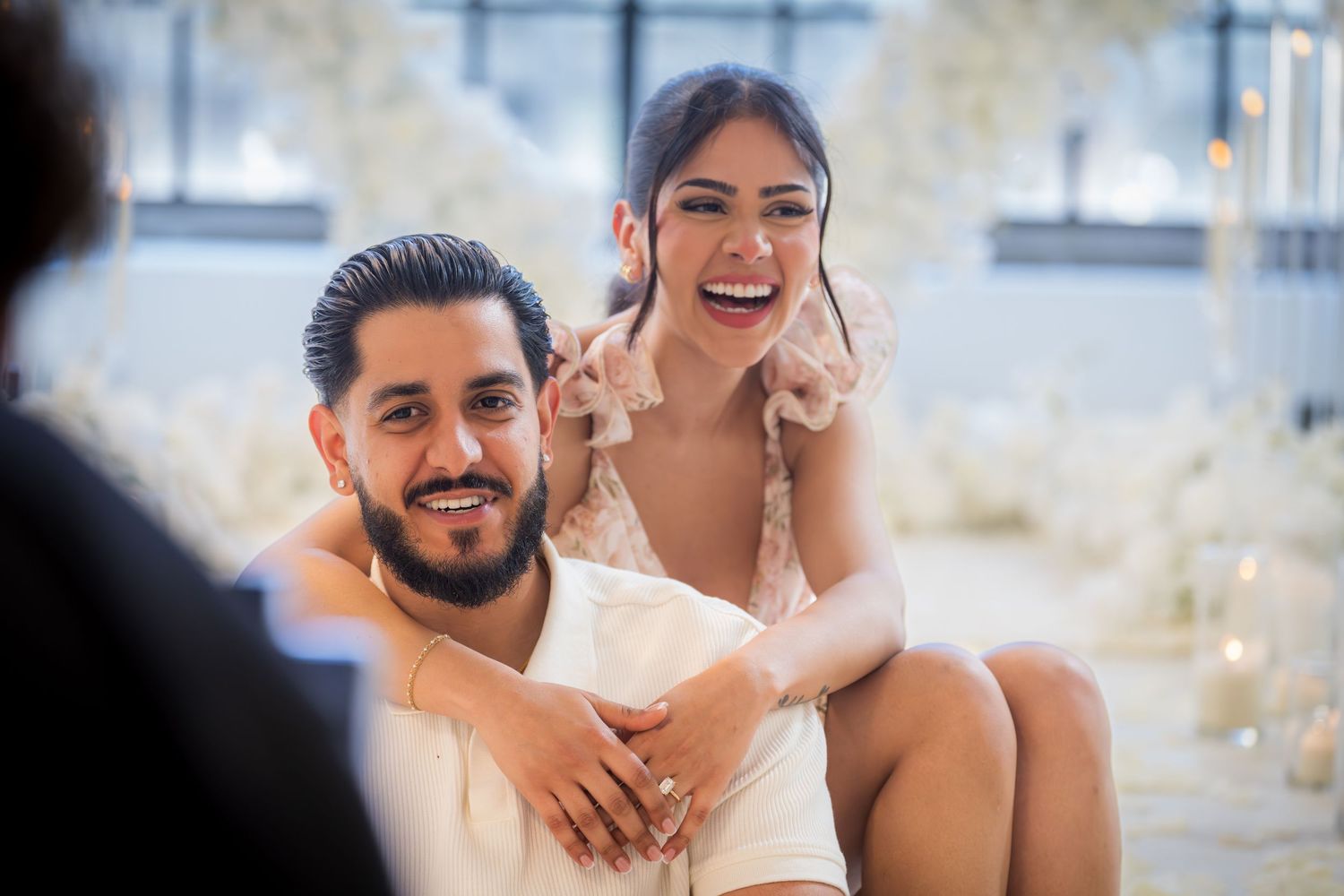 Happy couple laughing together, woman sitting on mans lap in elegant white floral decorated venue.
