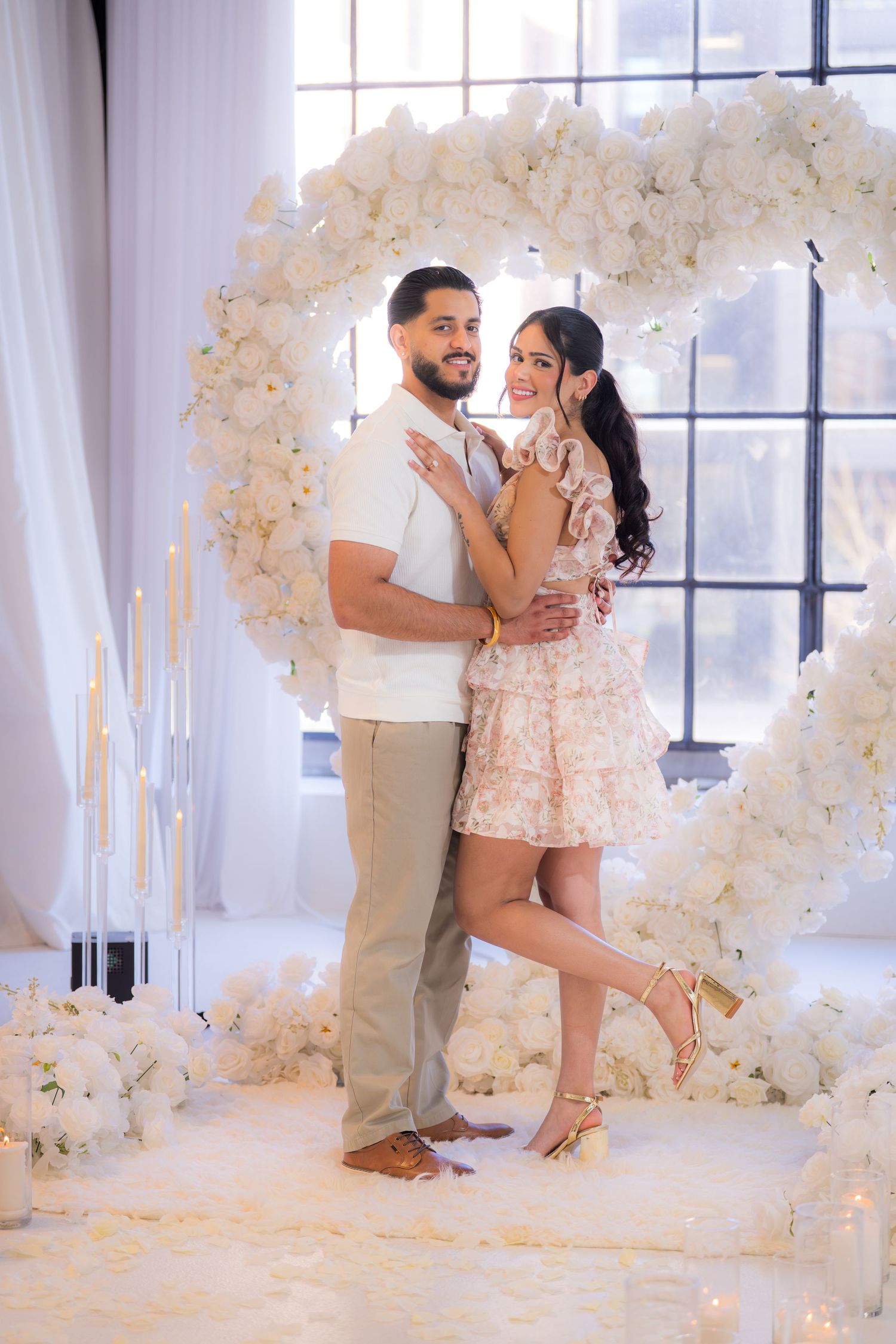Couple embracing in front of a white floral heart arch with candles in an elegant romantic setting.
