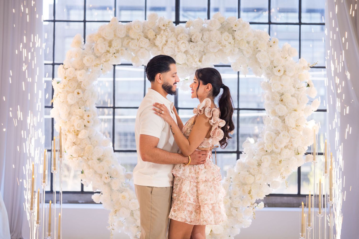 Romantic couple embracing under a white heart-shaped floral arch with sparkling lights.