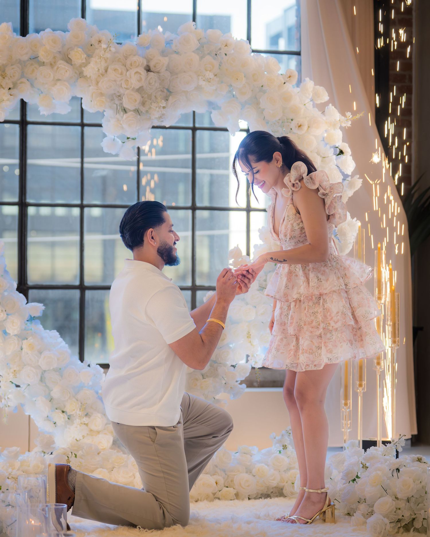 Man kneeling on one knee proposing to woman in floral dress under white flower arch.