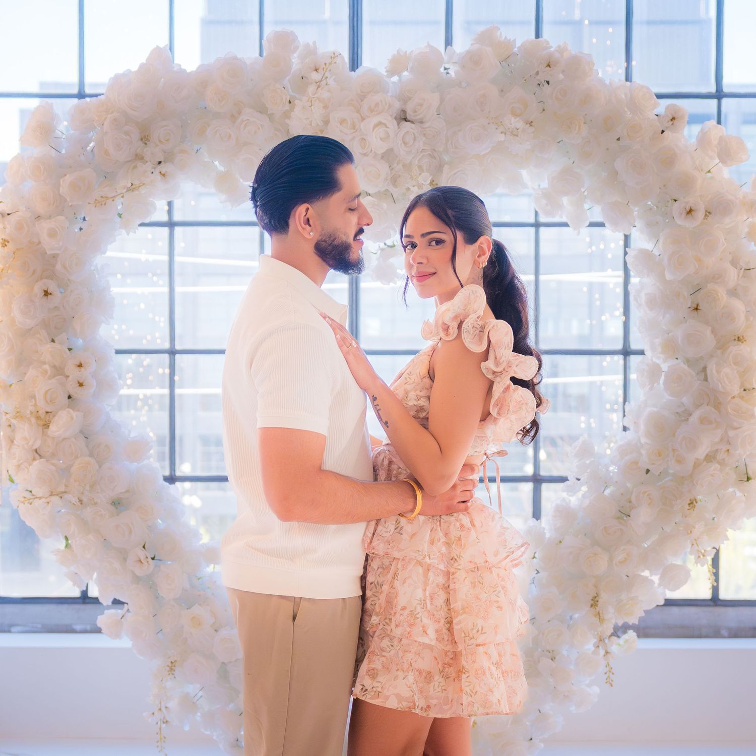 Engaged couple poses together in front of a stunning white flower heart arch in a bright venue.