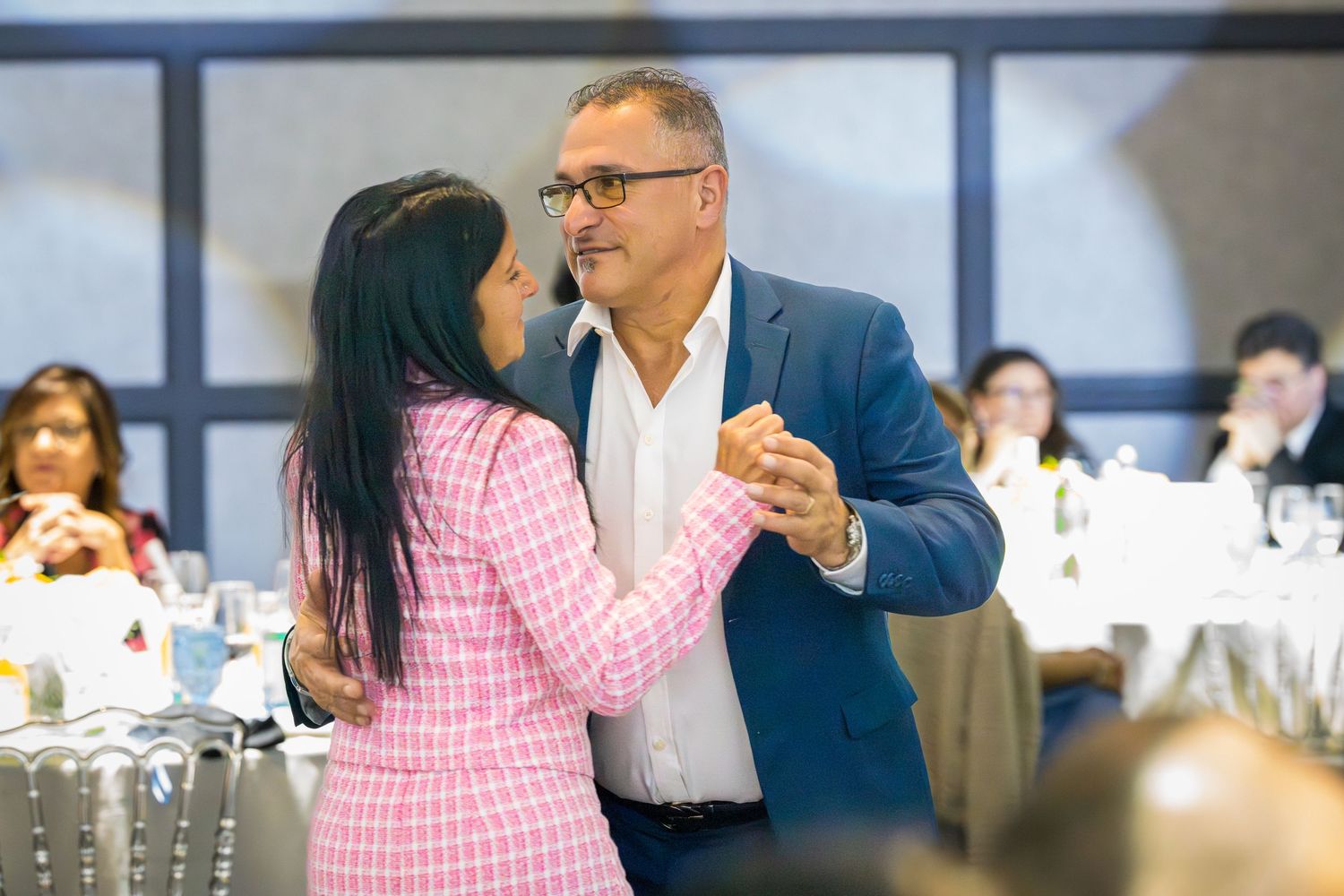 Couple dancing together at a formal banquet event, man in blue blazer and woman in pink tweed suit.