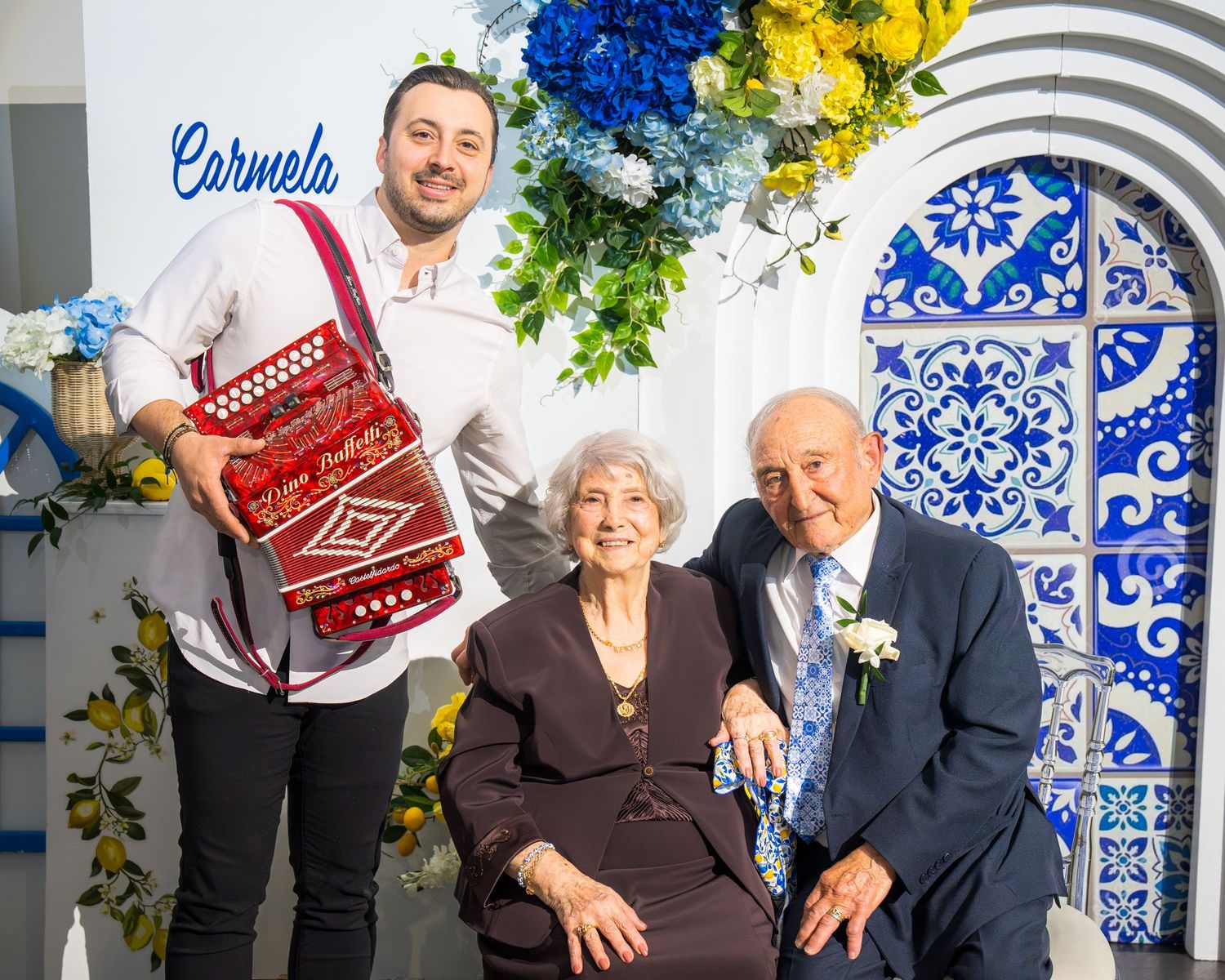 Young accordion player posing with smiling elderly couple before decorative blue Talavera tile backdrop at Carmela event.