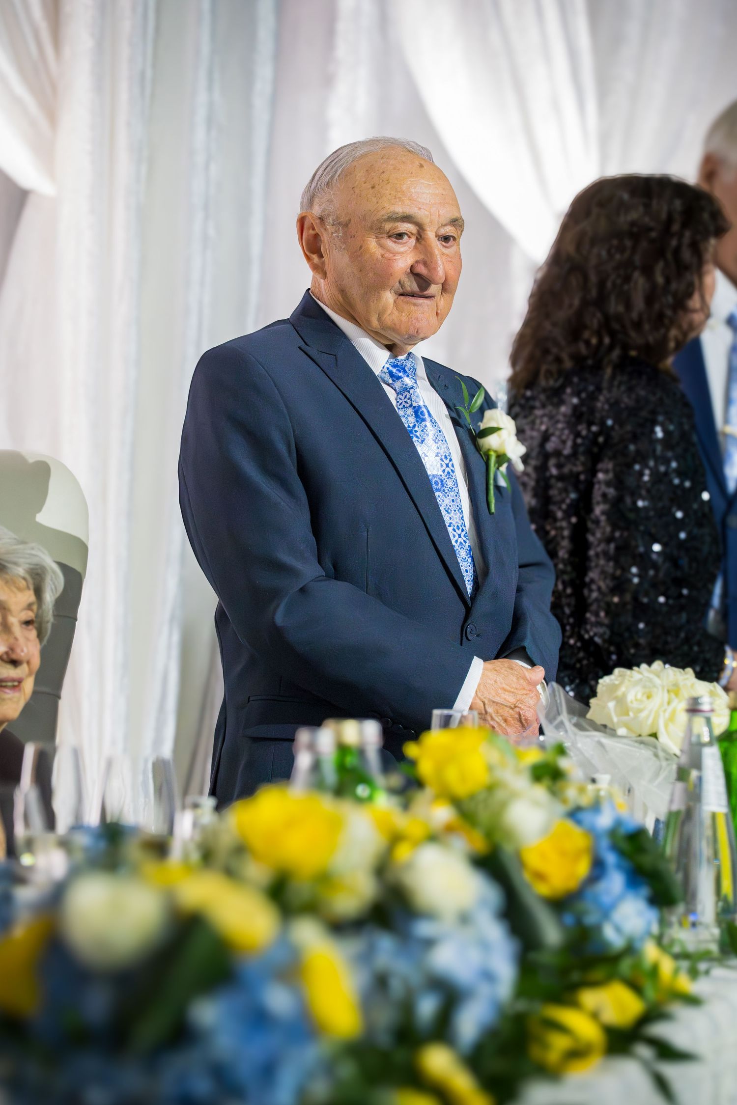 Elderly man in navy suit with blue floral tie and boutonniere smiling at a formal wedding reception.