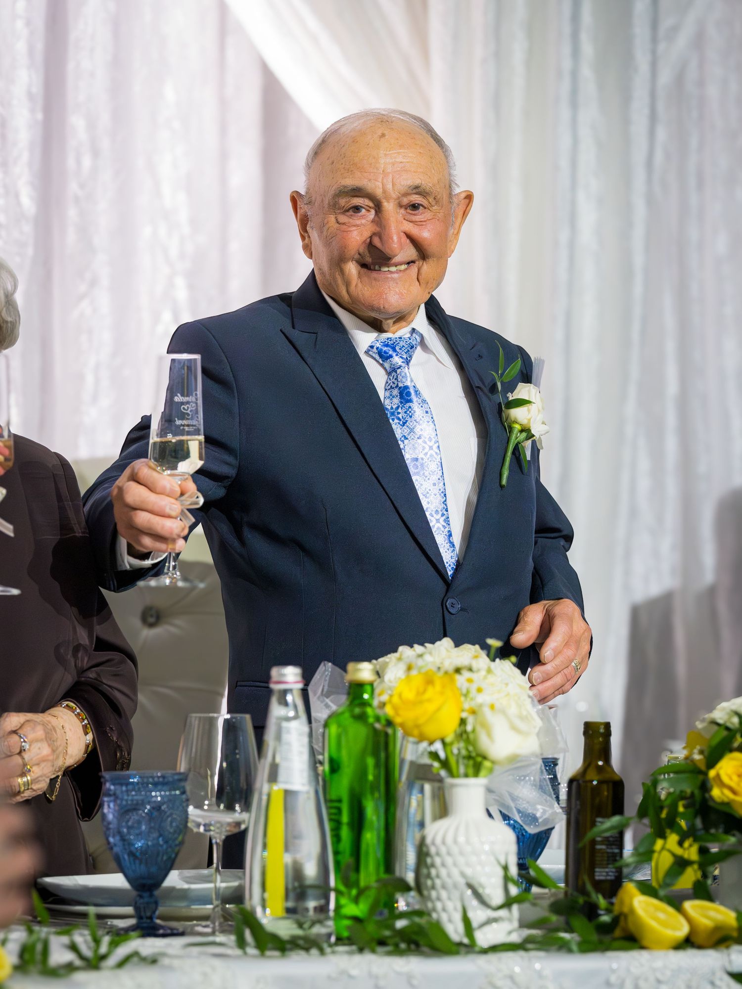 Elderly man in navy suit with blue tie raising champagne glass at 65th wedding anniversary celebration.