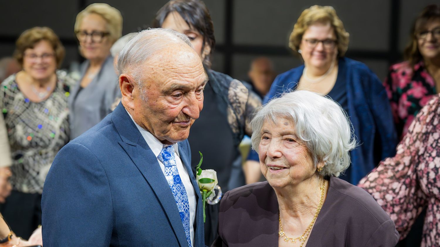Elderly couple sharing a tender moment together at a wedding celebration with guests in background.