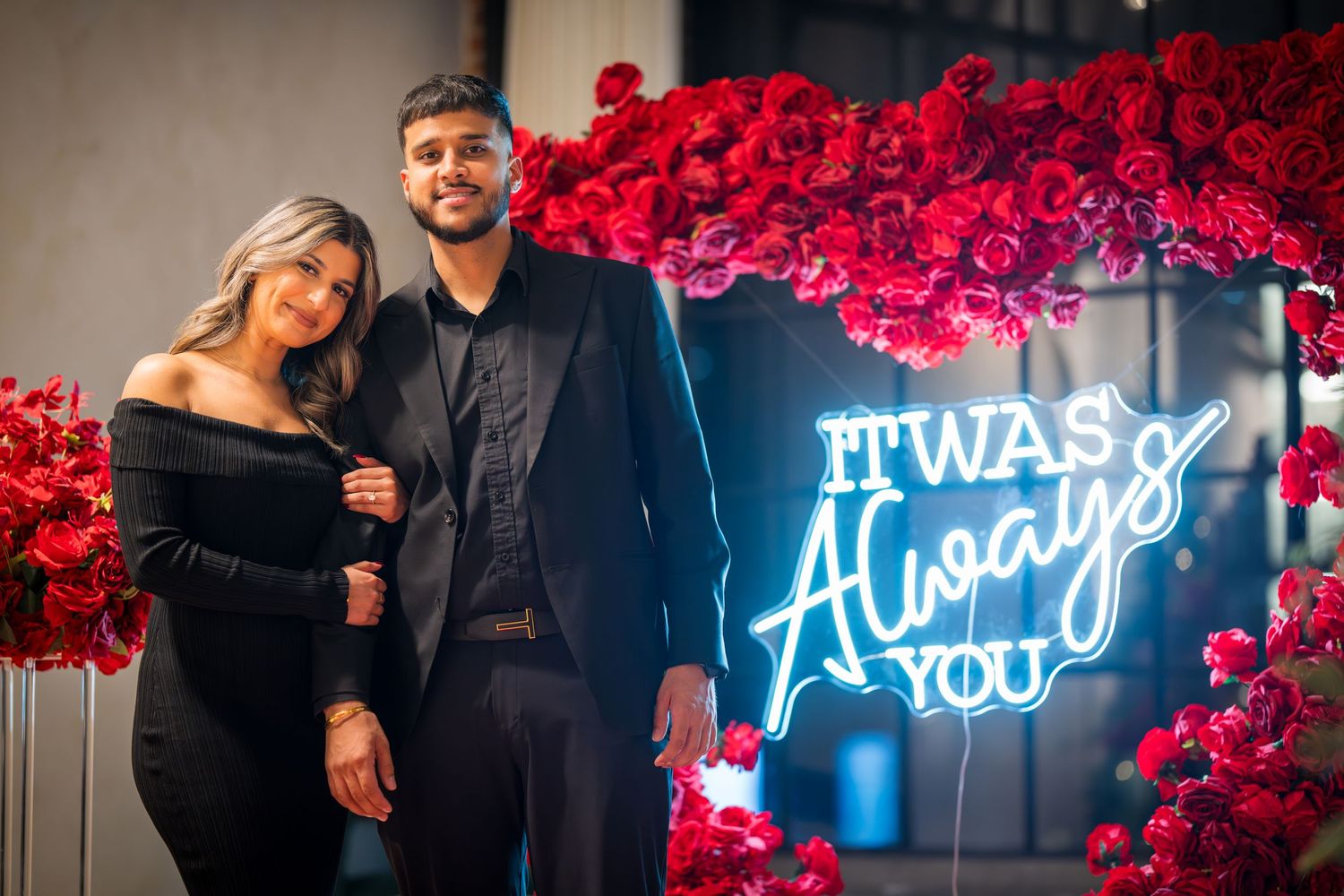 Couple stands before a neon sign reading 'It Was Always You' surrounded by red roses.