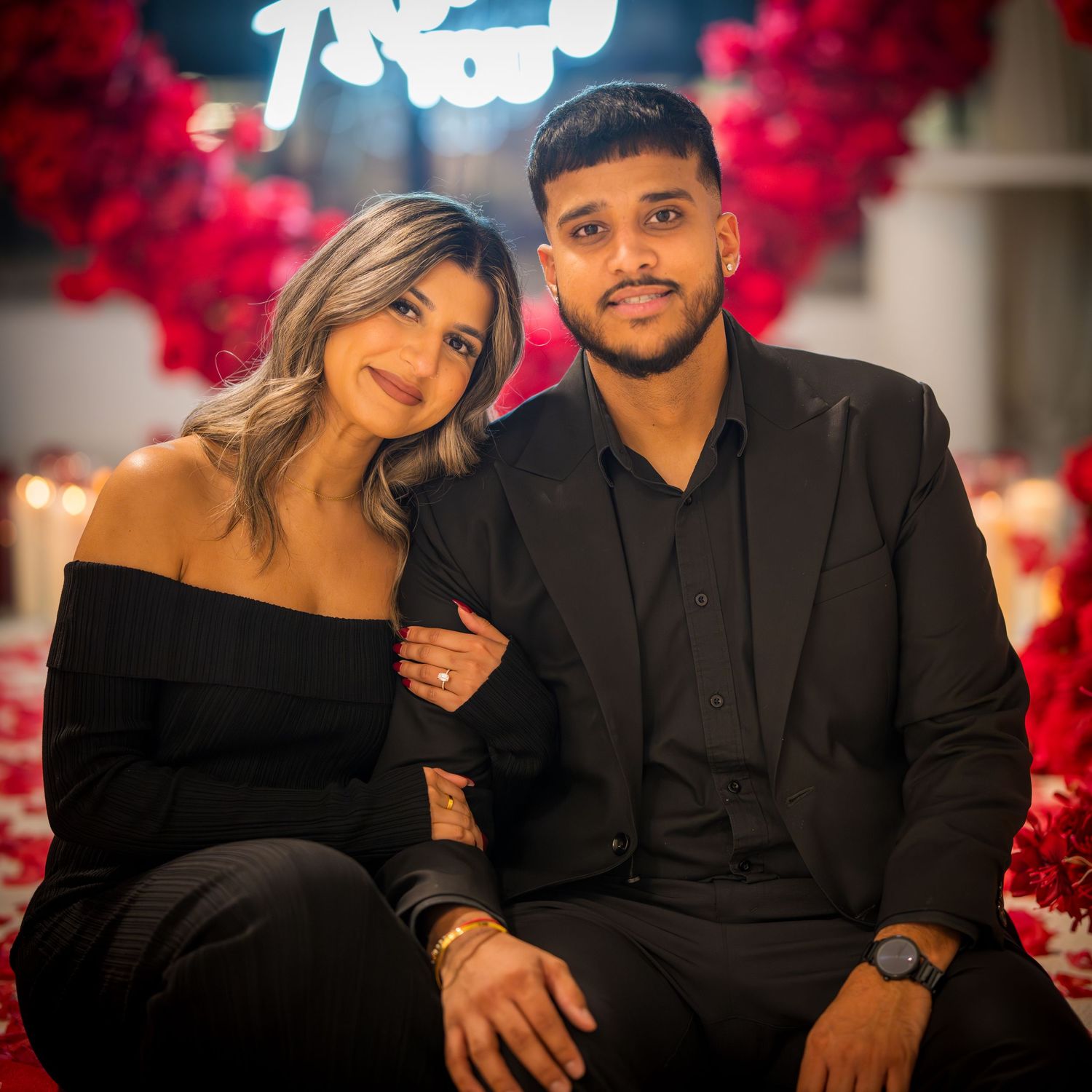 Couple in coordinated black outfits poses against red rose backdrop with neon lighting.