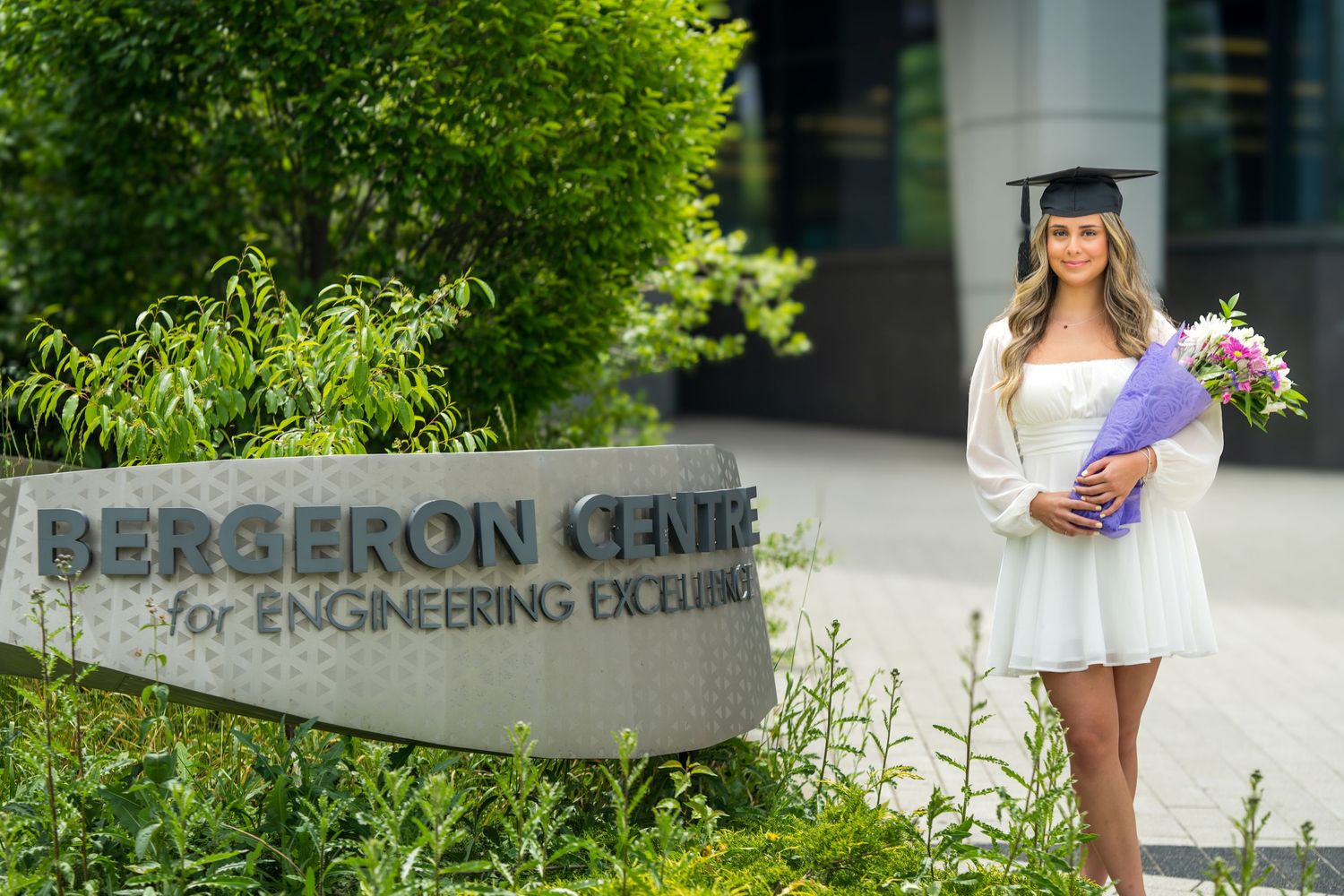 Graduate in white dress and cap holding purple flowers stands outside Bergeron Centre building.