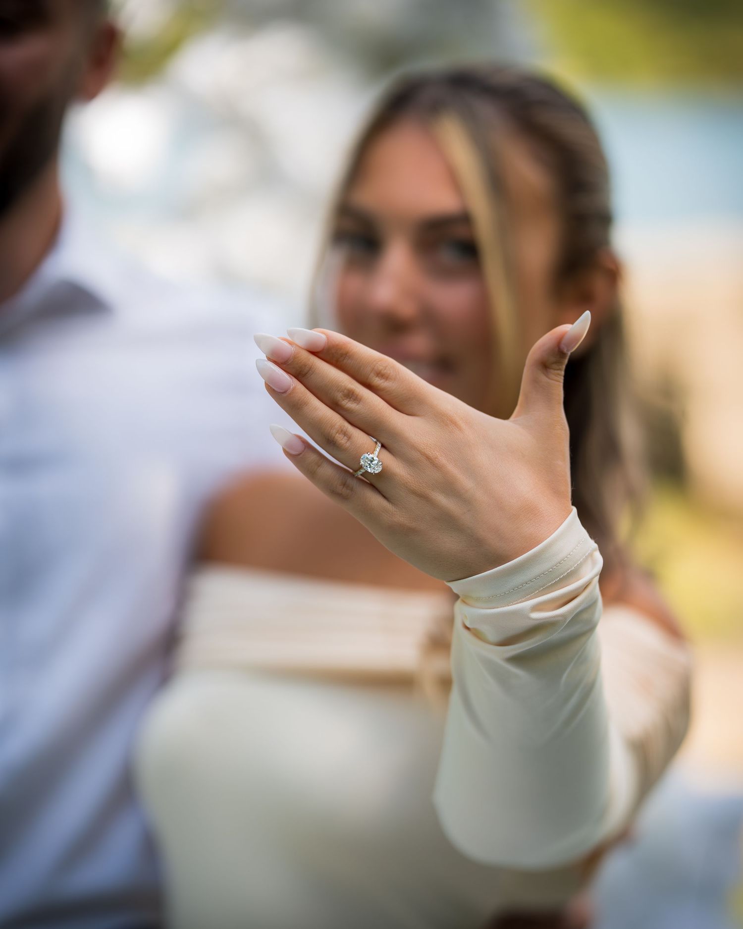 A hand displaying a diamond engagement ring with an off-shoulder white dress sleeve.