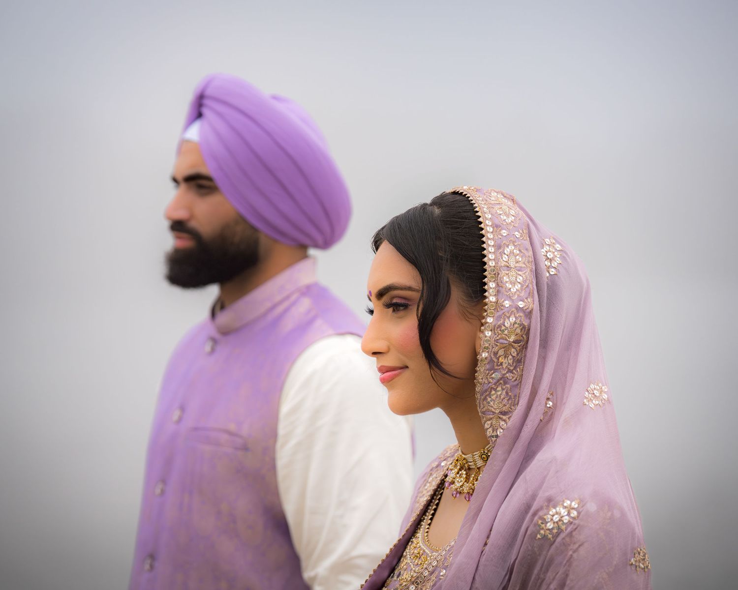 A couple in elegant purple and white traditional Indian attire poses against a misty background.