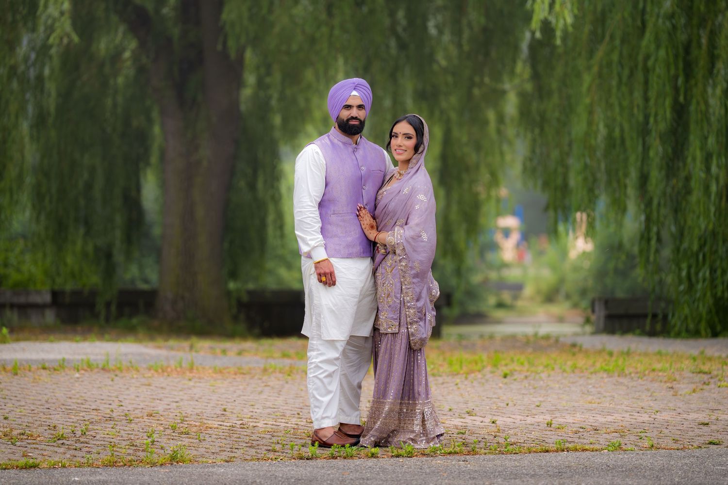 A couple in coordinating lavender and white traditional Punjabi outfits pose under a weeping willow tree.