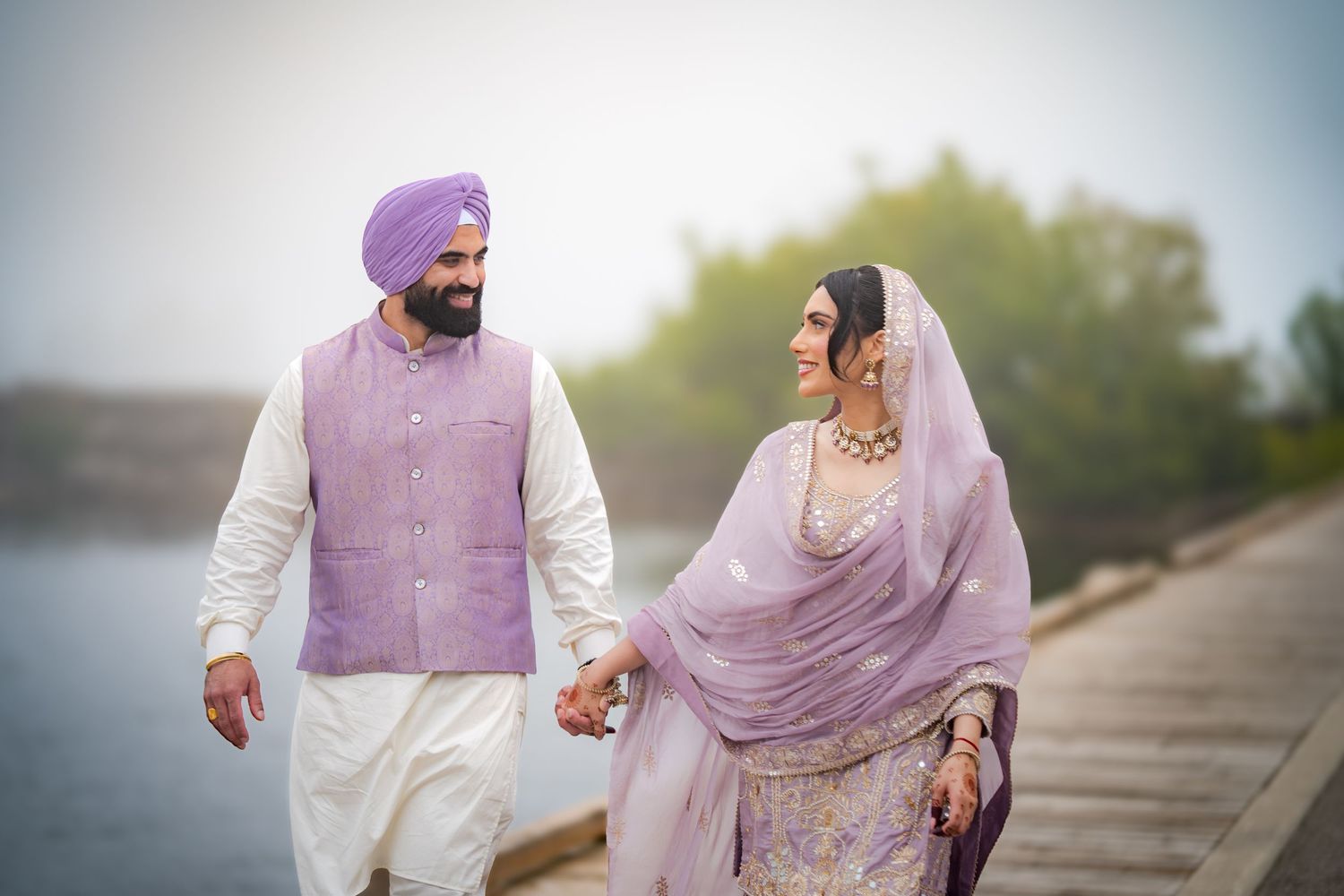 A couple in coordinated lavender and white traditional Indian wedding attire pose outdoors in soft lighting.