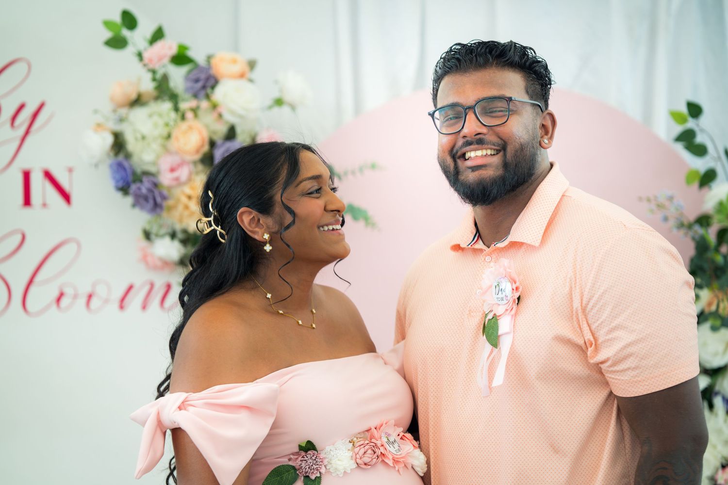 Couple poses in front of floral wall at pink-themed celebration event.