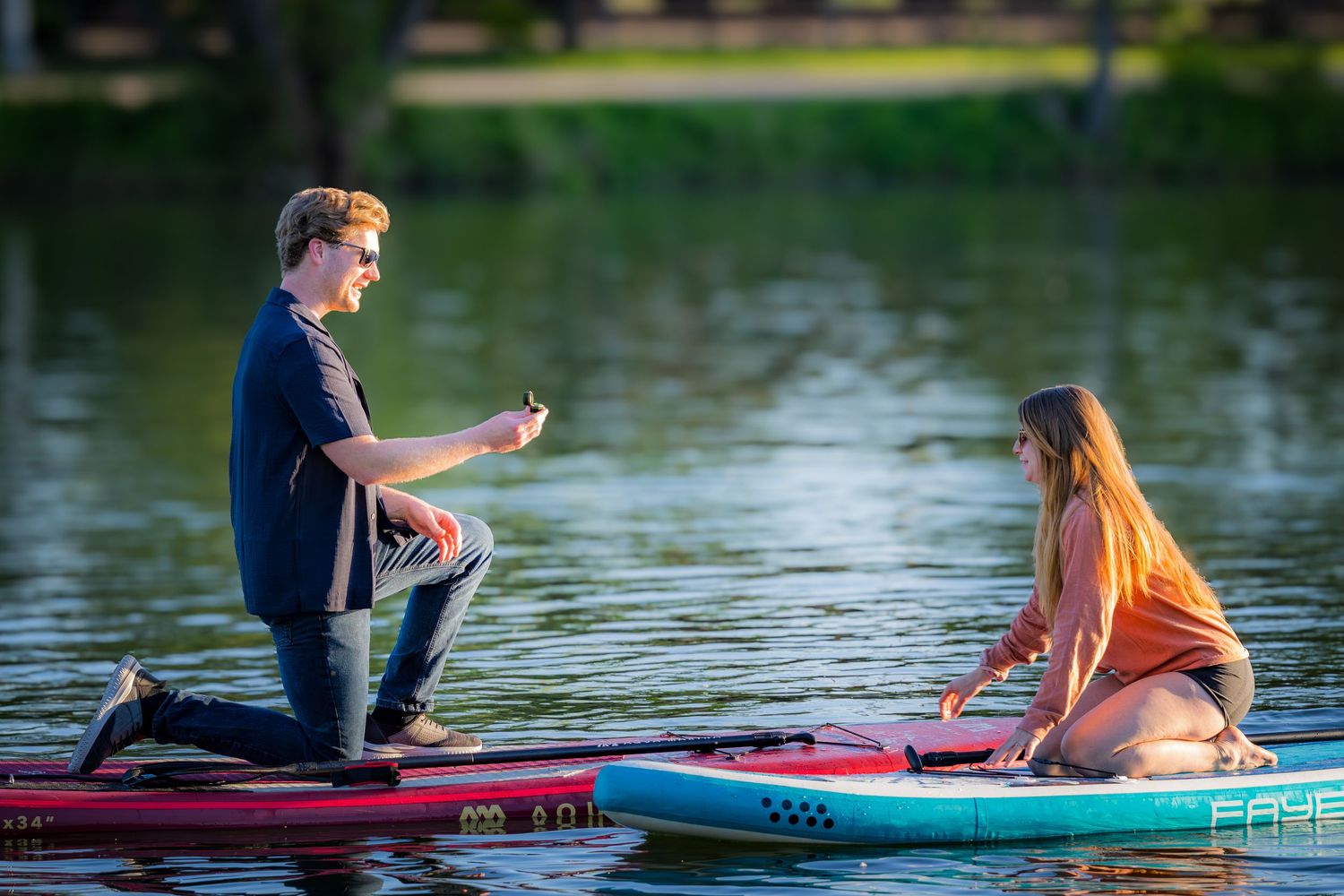 Man kneeling on a paddleboard proposing to a woman on a separate board on a sunny lake.