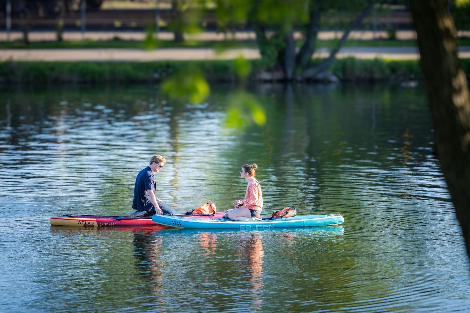 Two people sitting on paddleboards on a calm, sunlit lake surrounded by lush green trees.