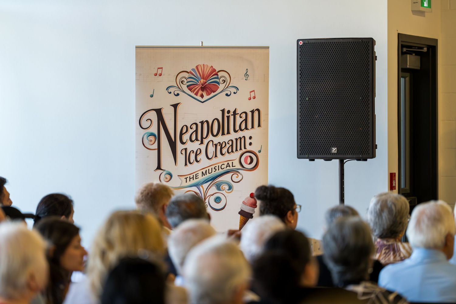 Audience seated before a Neapolitan Ice Cream The Musical banner and speaker at an indoor performance venue.