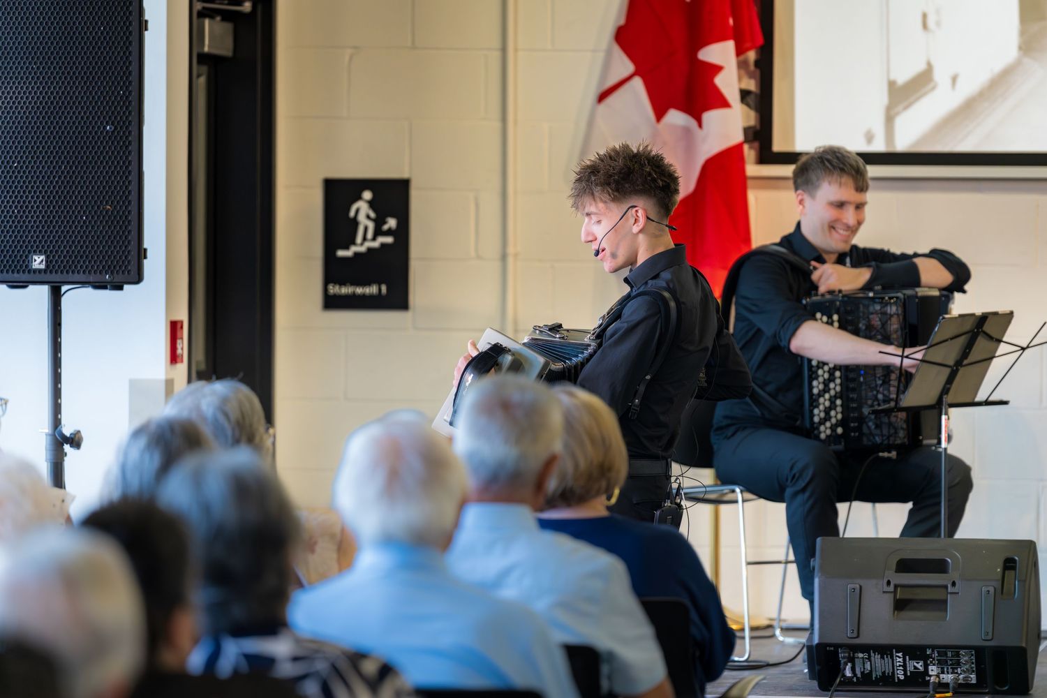 Two musicians performing violin and accordion for seated audience at Canadian cultural event.