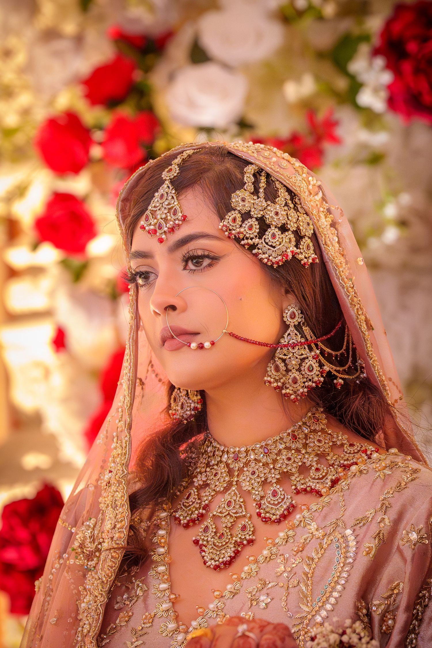 Bride in pink lehenga with detailed jewelry and makeup poses against floral backdrop with red roses.
