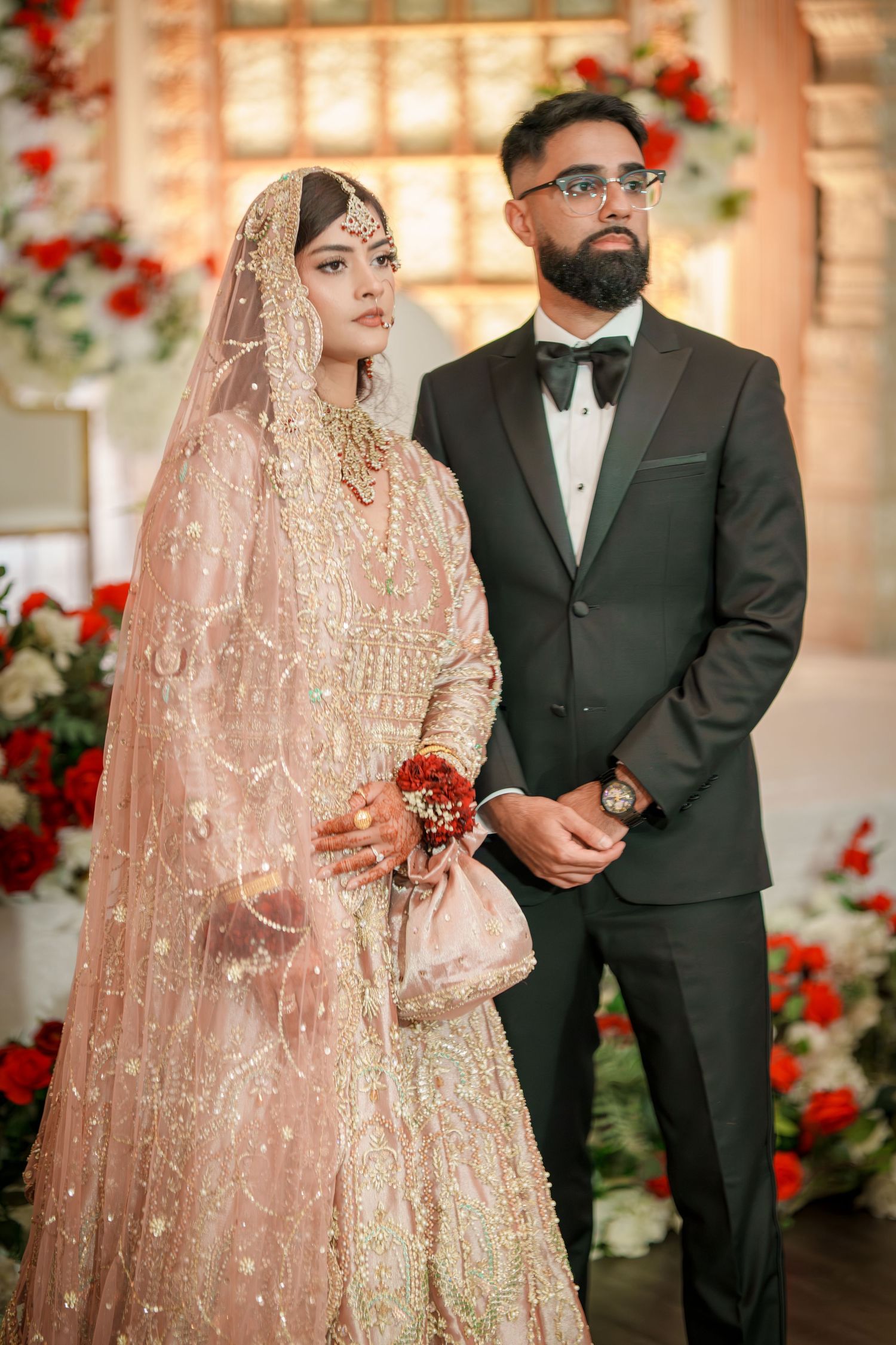 Elegant wedding couple in traditional South Asian attire pose against floral backdrop with red roses.
