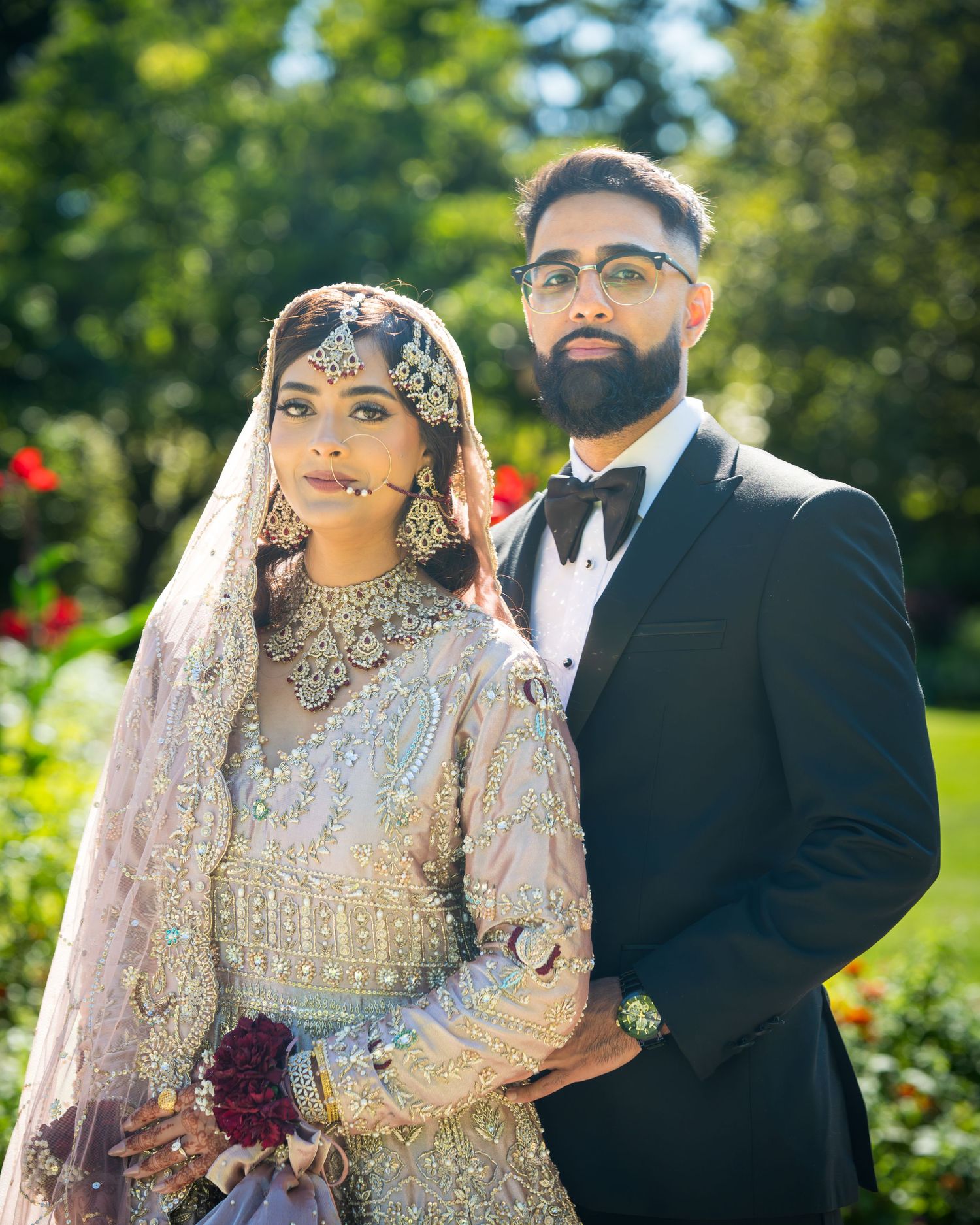 Traditional South Asian wedding couple poses in outdoor garden setting with vibrant greenery and floral backdrop.