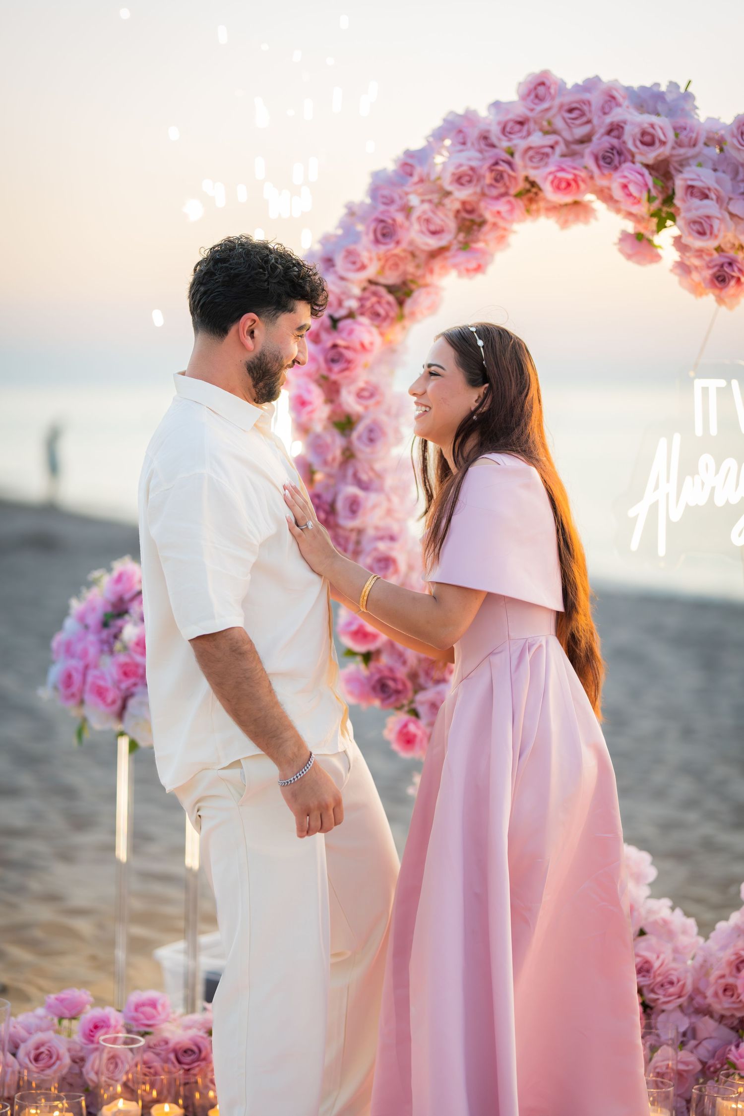 A couple shares an intimate moment under a pink floral arch on a beach at sunset.