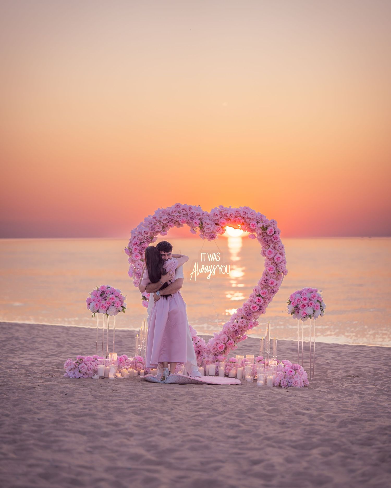 Heart-shaped floral arch with pink flowers on beach at sunset with glowing candles creating romantic ambiance.