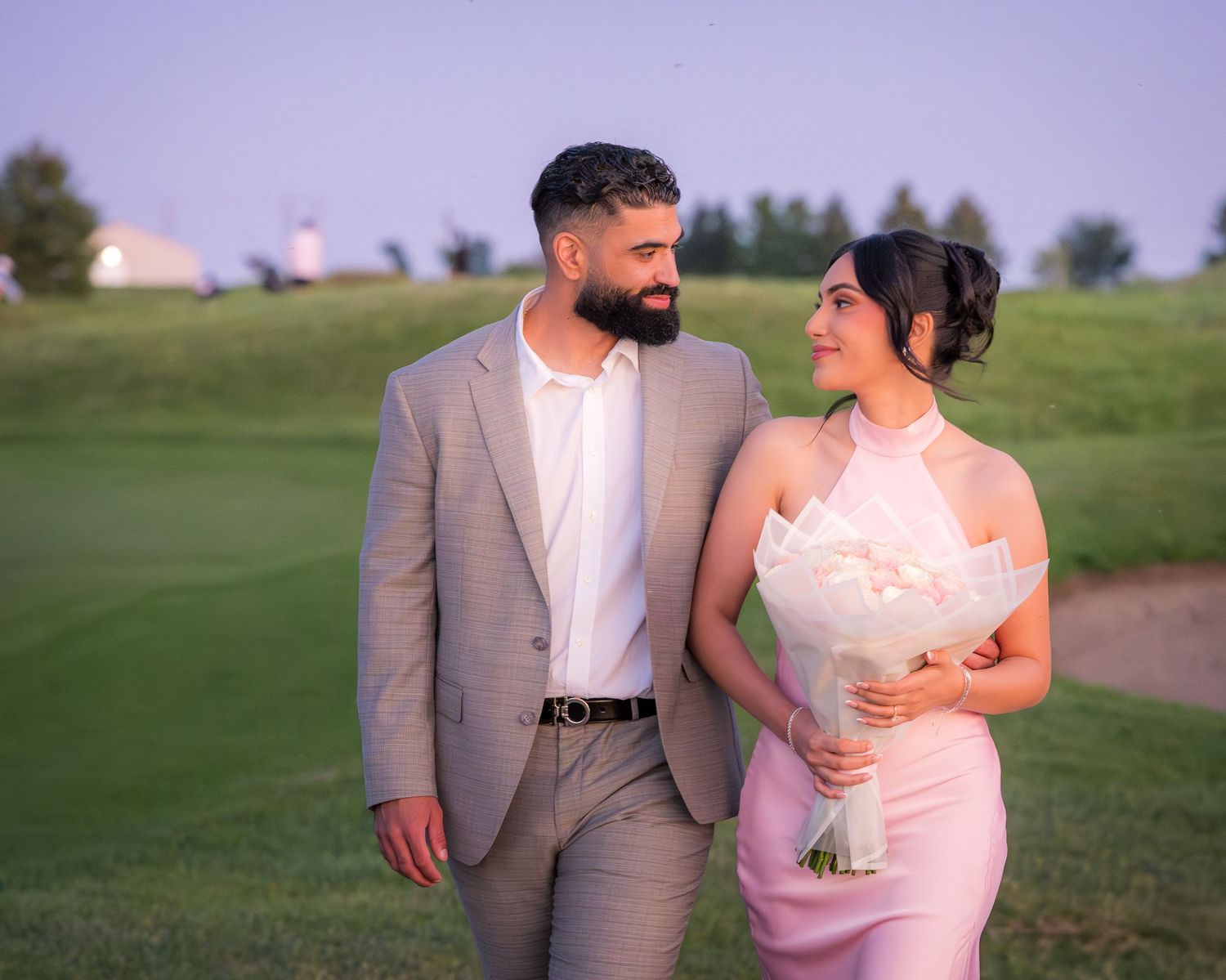 A couple in formal attire poses together on a grassy path during golden hour.