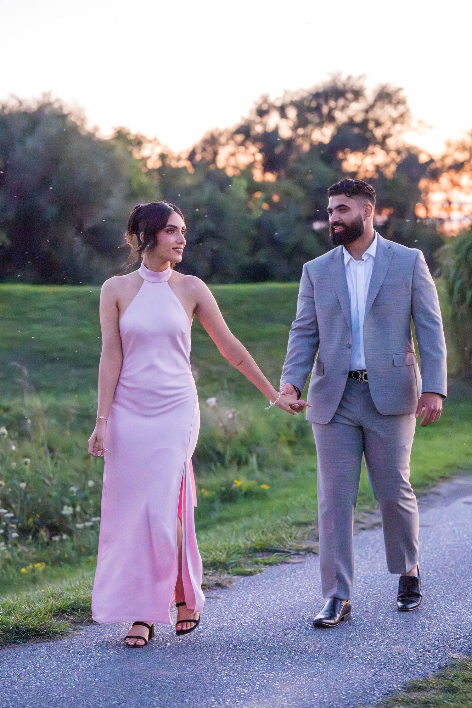 Two people in formal wear walk hand in hand on a gravel path during golden hour.