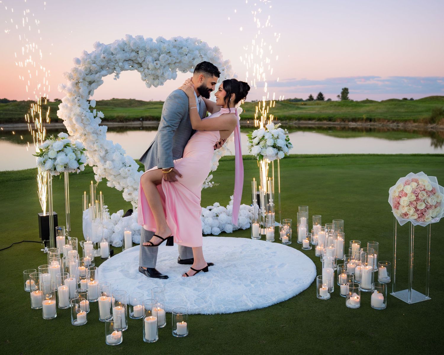 Sunset wedding ceremony setup with white floral arch and glowing lanterns on manicured golf course.
