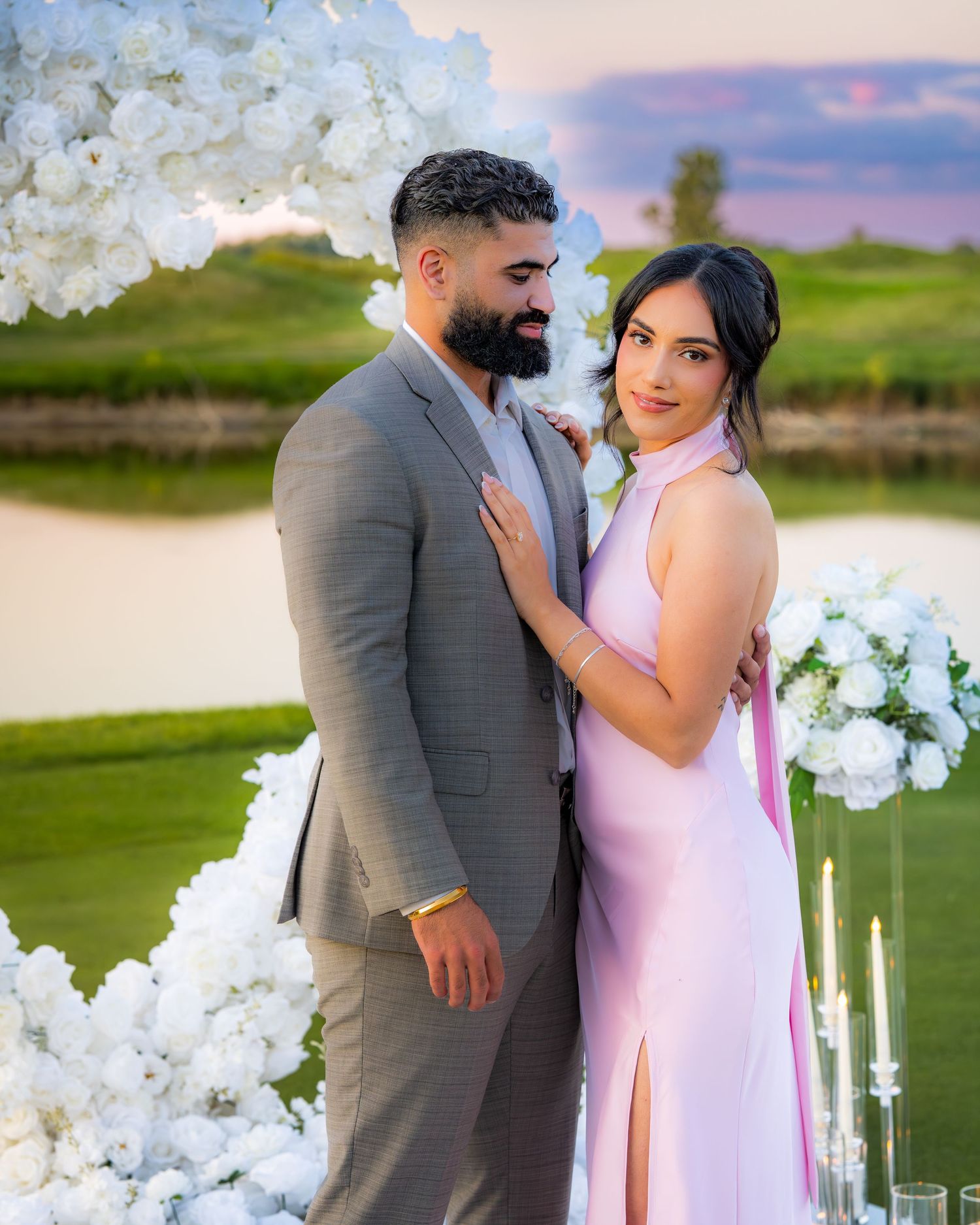 A couple shares a romantic moment on a golf course surrounded by white flowers and candles.