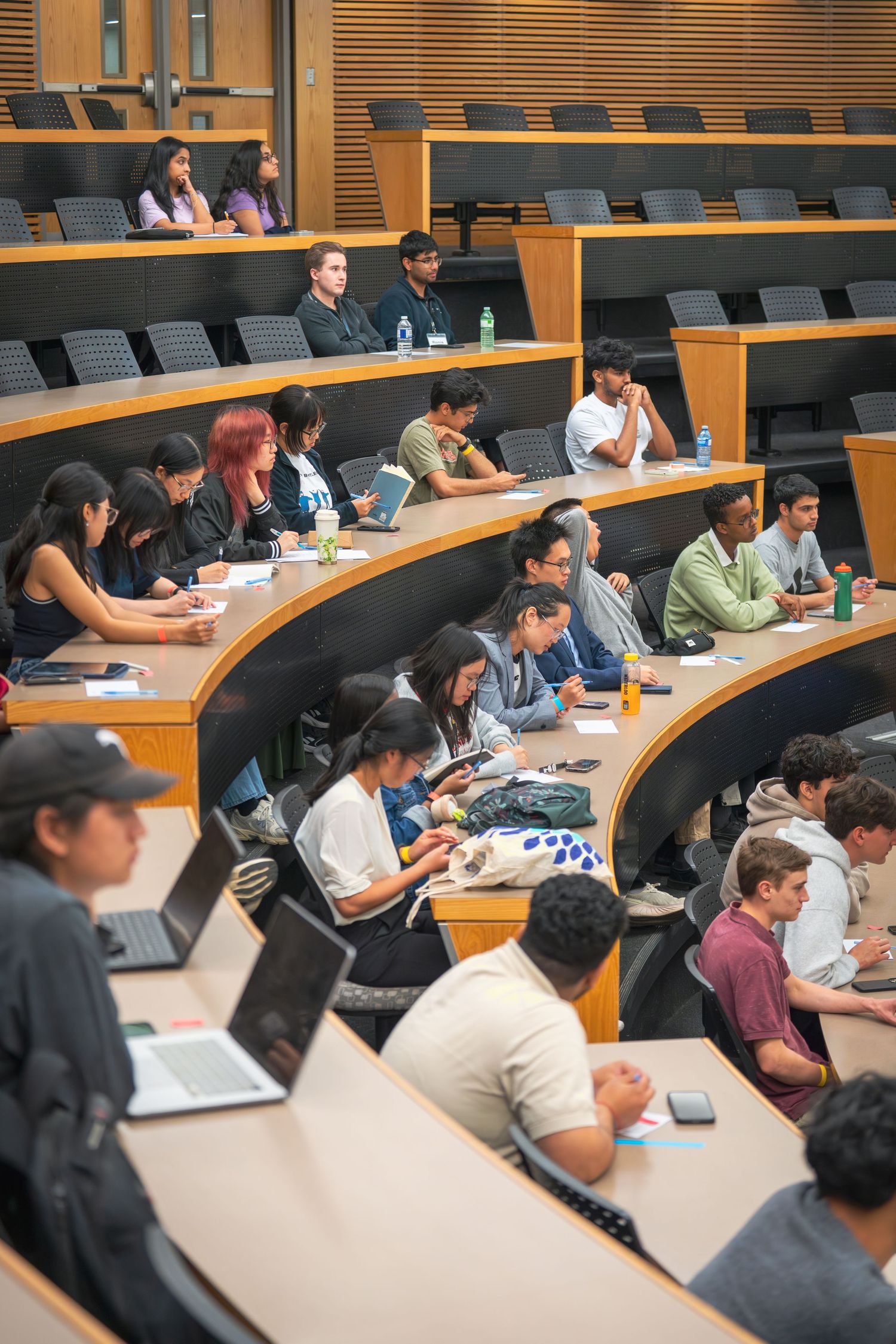 Students seated in tiered lecture hall rows taking notes during a presentation.