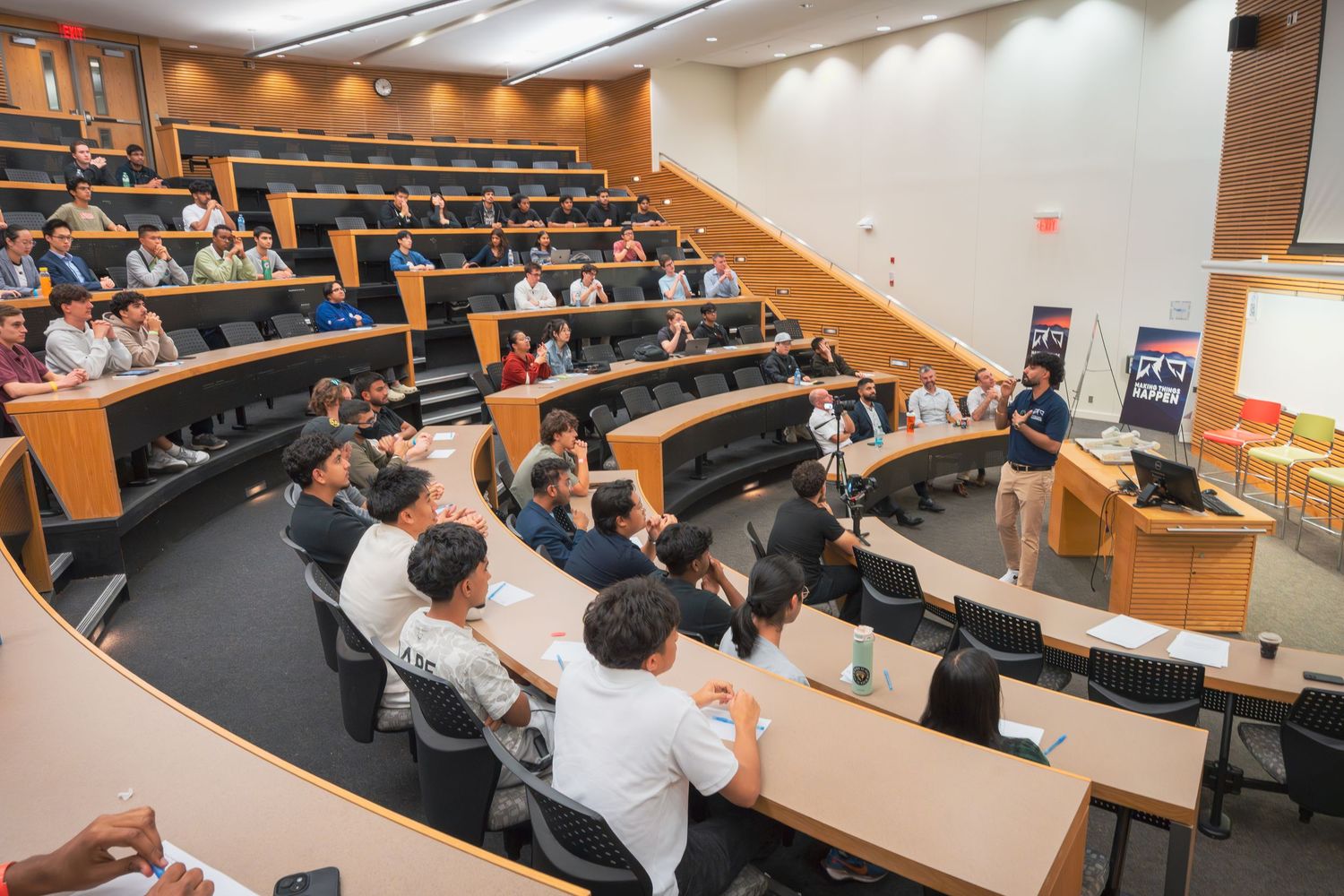 People seated in a modern lecture hall with wooden paneled walls and tiered seating during a presentation.