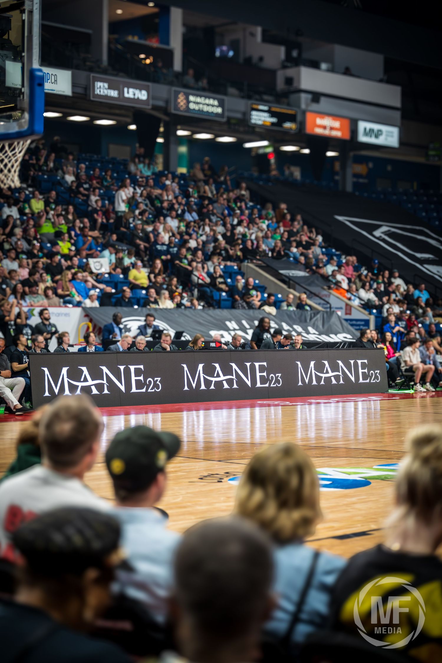 Basketball arena crowd with courtside MANE23 banners displayed during a professional game.