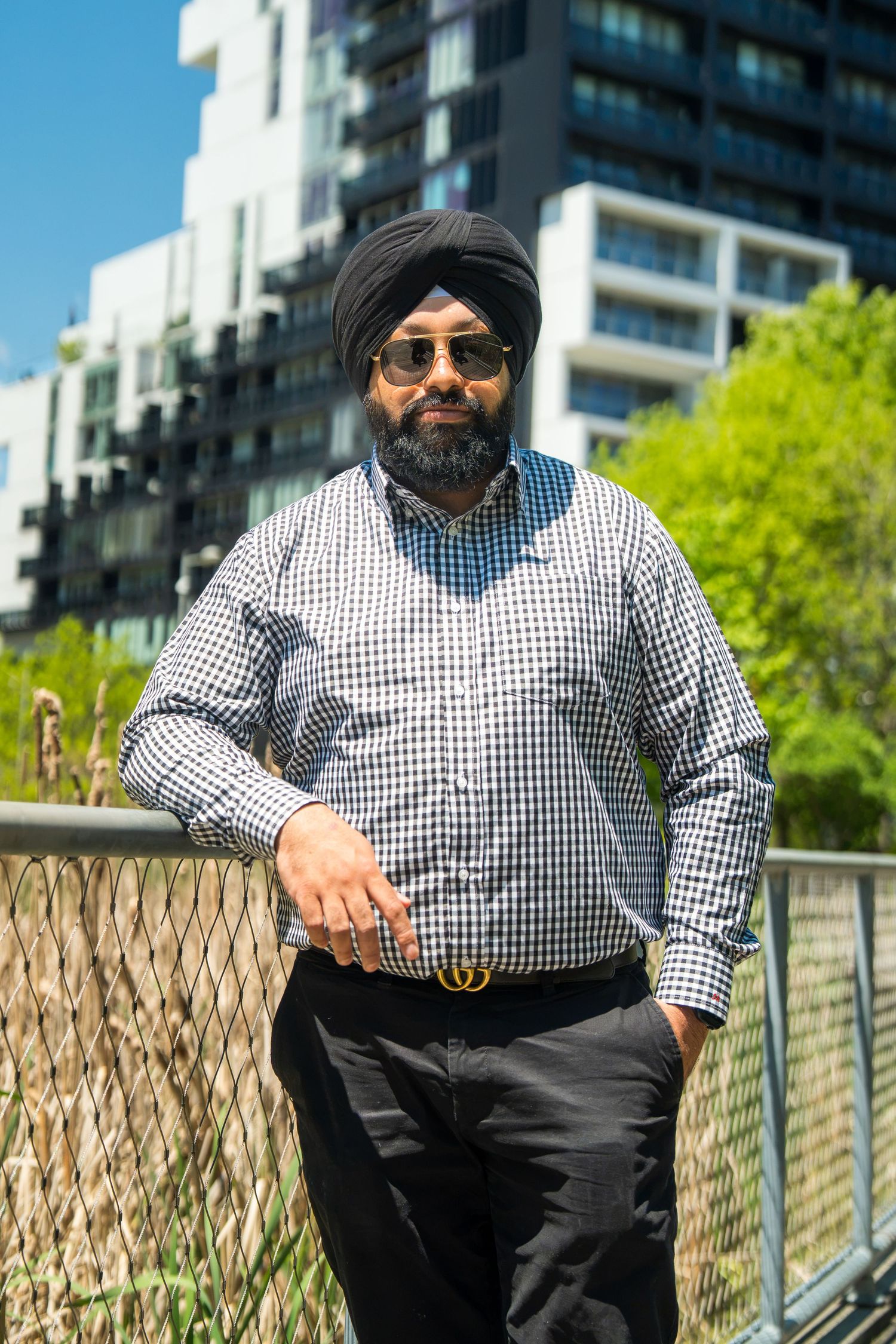 Sikh man in black turban and plaid shirt leaning on fence with modern buildings in background.