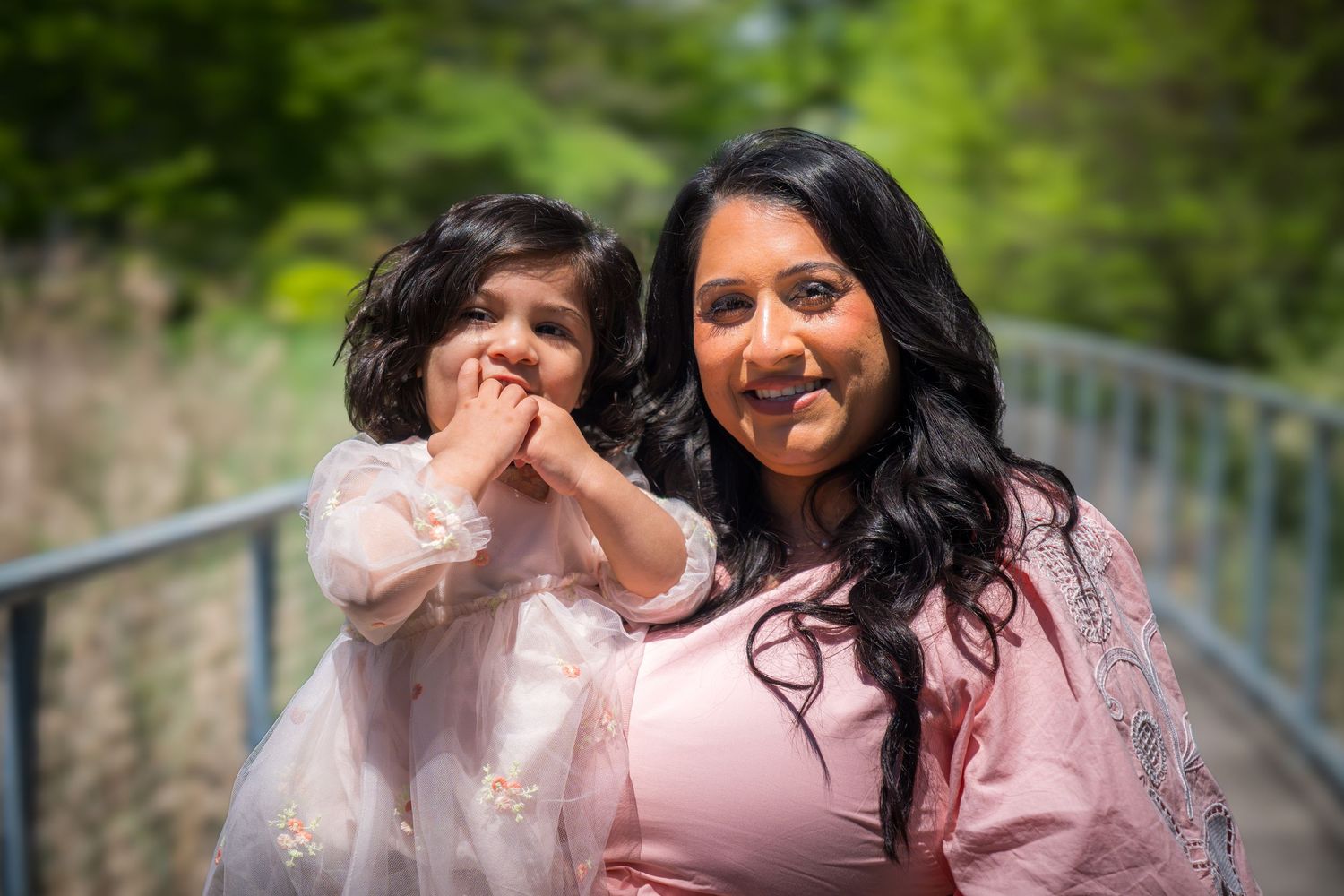 A woman and young girl in matching pink outfits pose together on a bridge surrounded by green trees.