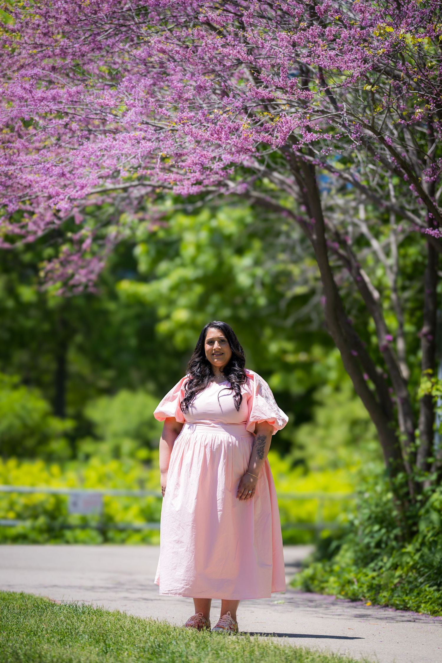 Pregnant woman in pink dress stands under blooming pink redbud tree in spring park.