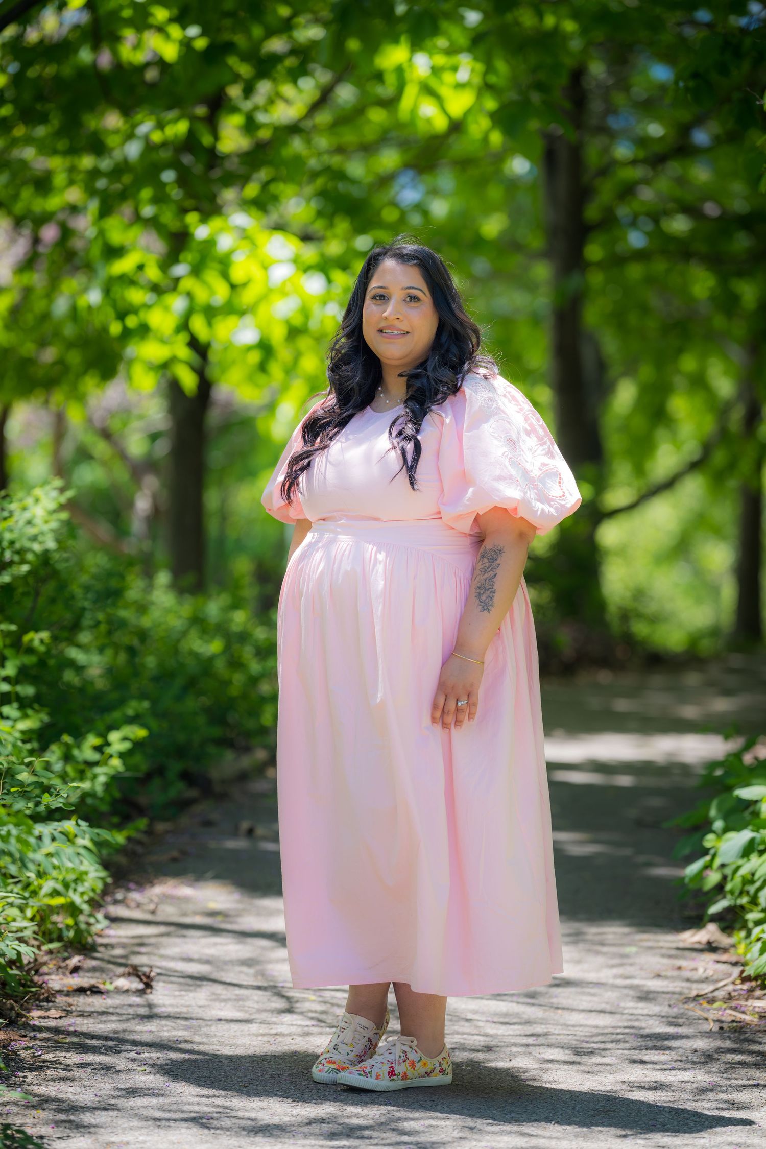 Smiling pregnant woman in pink midi dress with puff sleeves poses on sunlit park path.