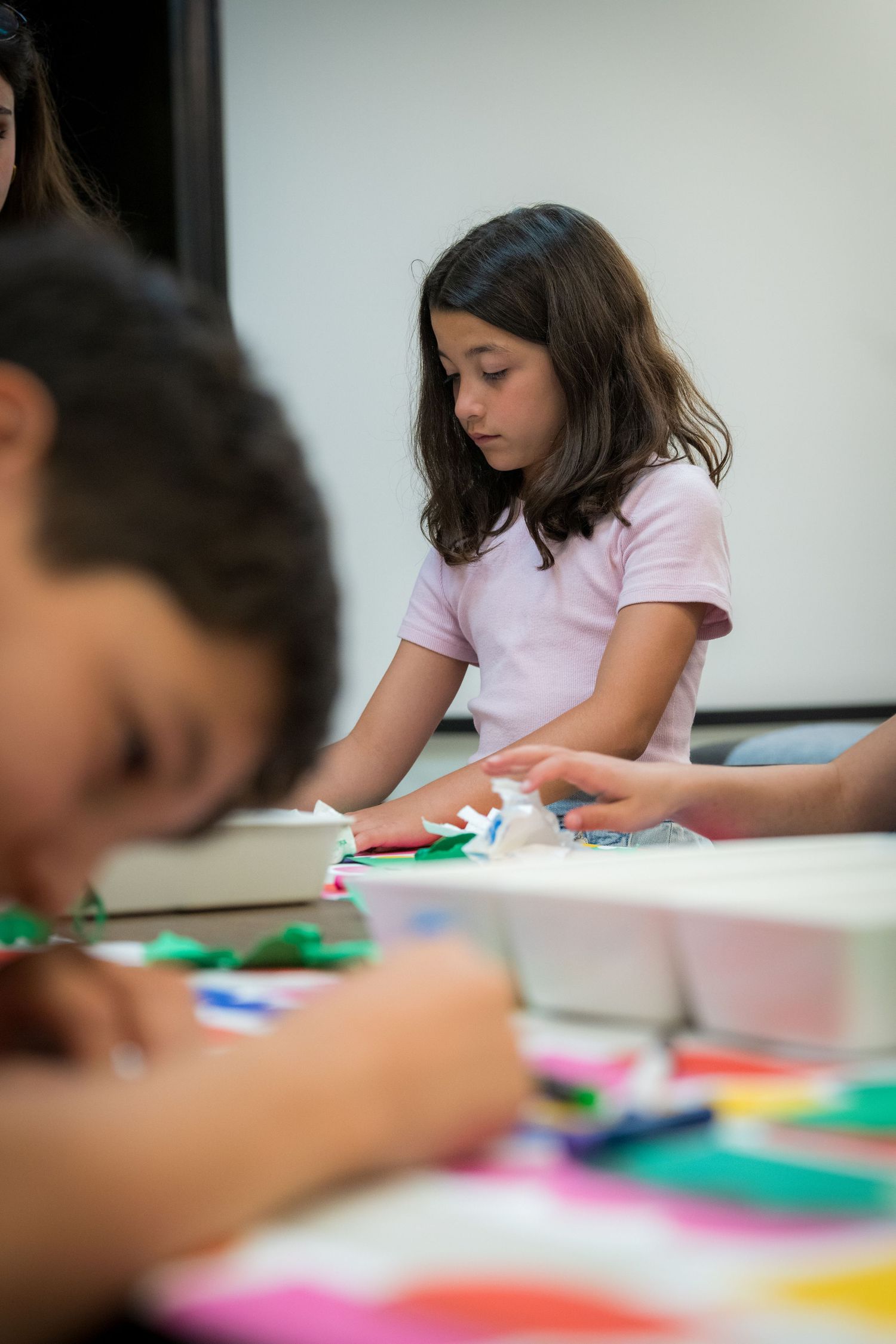 A girl in a pink shirt concentrates on a colorful paper craft activity at a table.