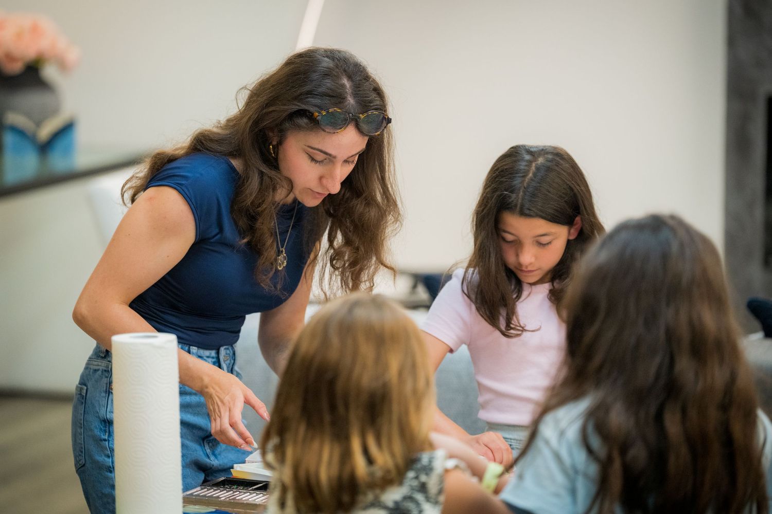 A woman teacher engages with three young girls during a creative art activity at a bright indoor classroom table.