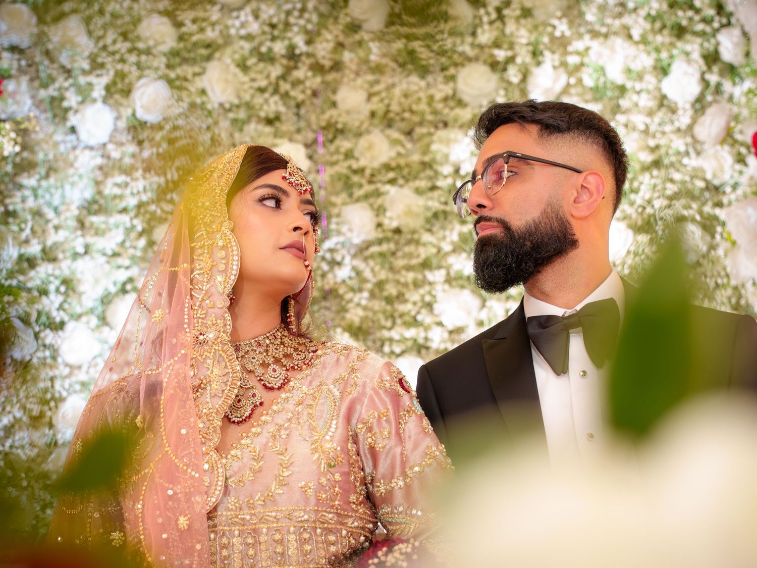 Wedding couple poses under floral arch with soft bokeh lighting effects.