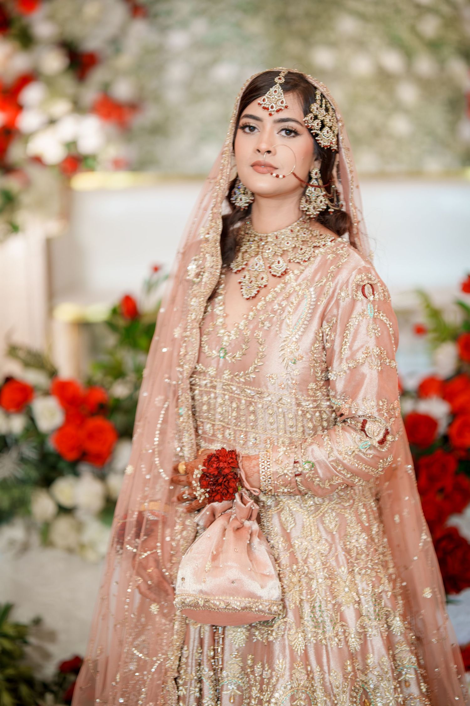 Traditional South Asian bride in ornate pink lehenga with jewelry and floral backdrop.