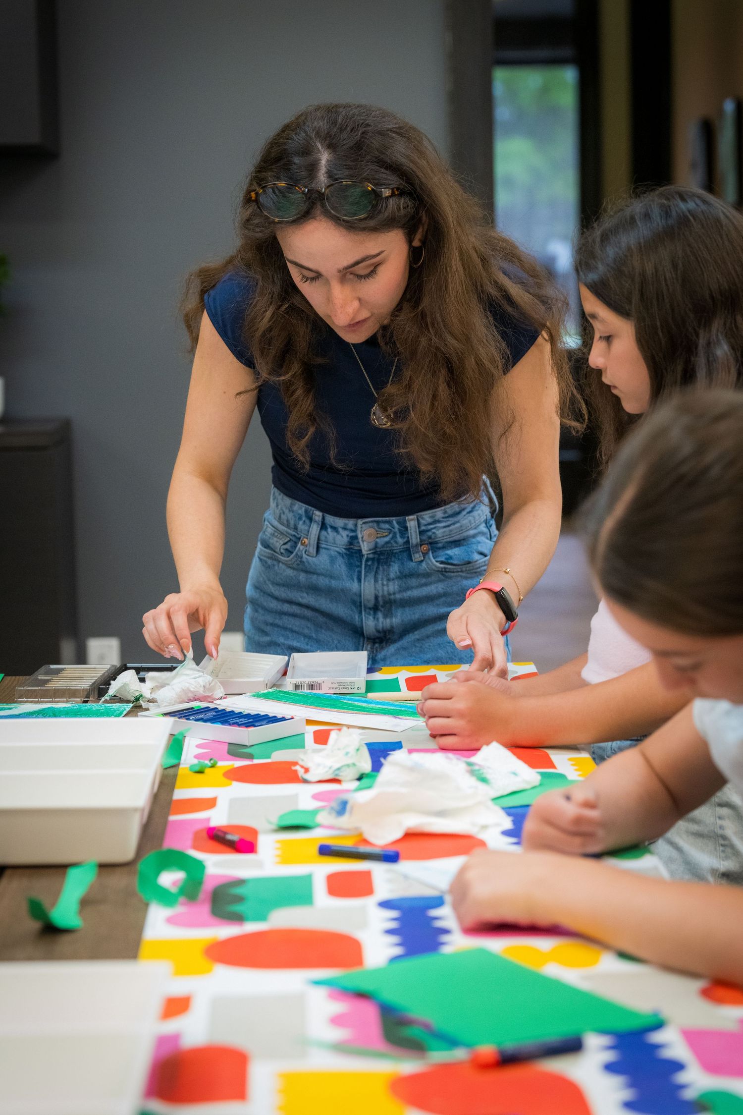 Woman in blue top guides two children in a colorful paper craft activity at a table.