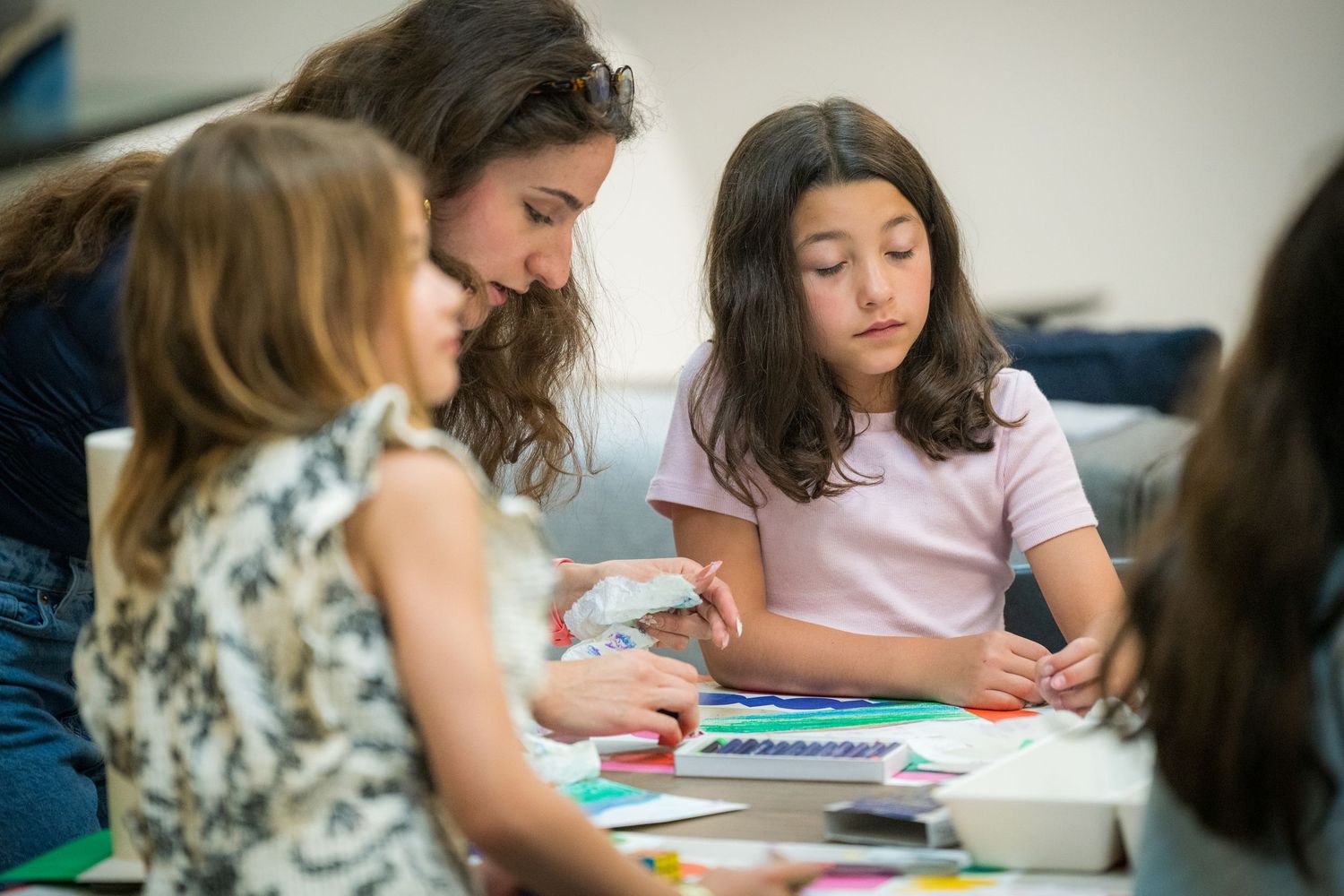 Female art teacher assists two young girls with a colorful craft project at a classroom table.