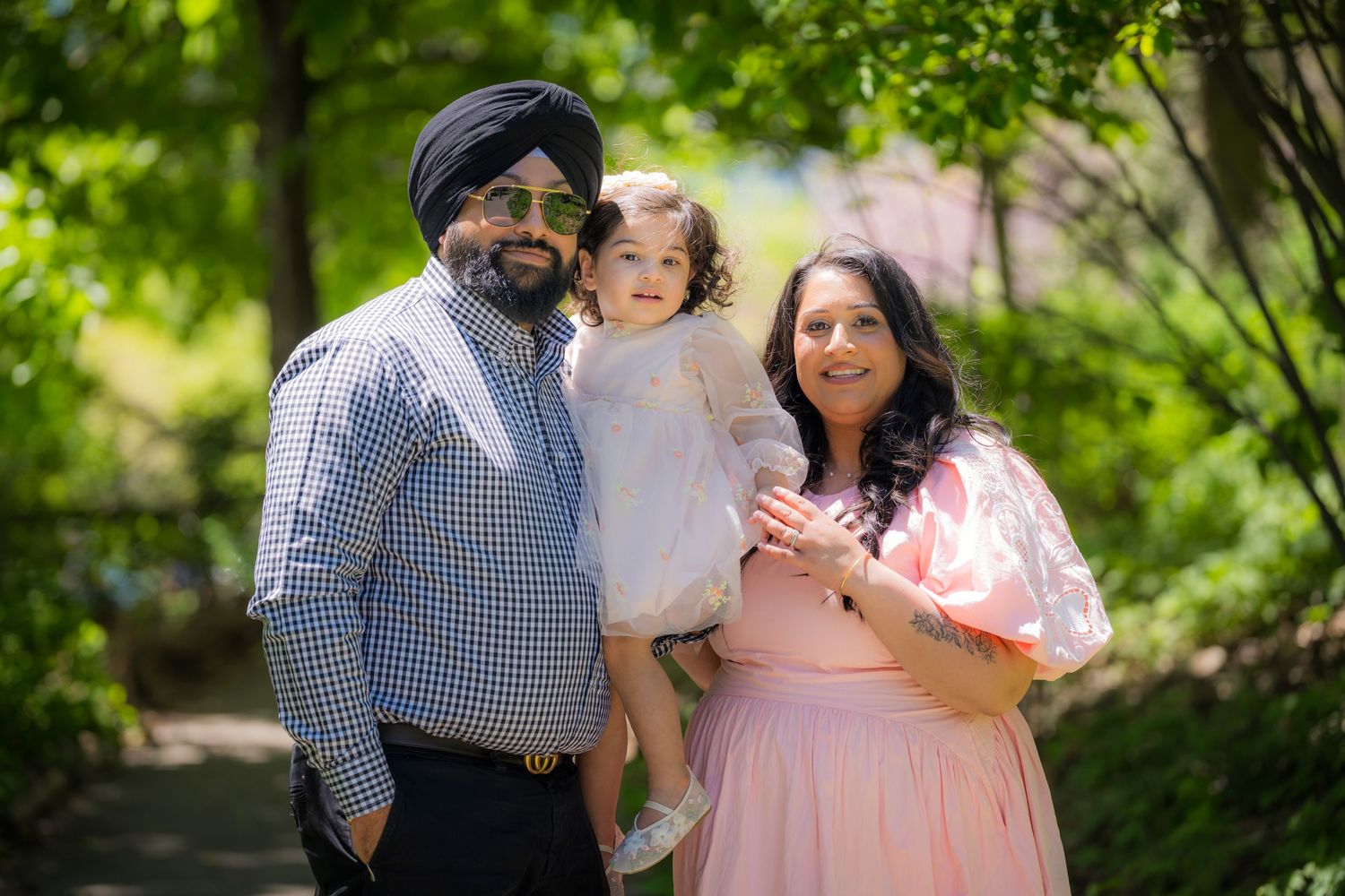 A Sikh family of three smiling outdoors, father in turban holding toddler, mother in pink dress, lush green trees.
