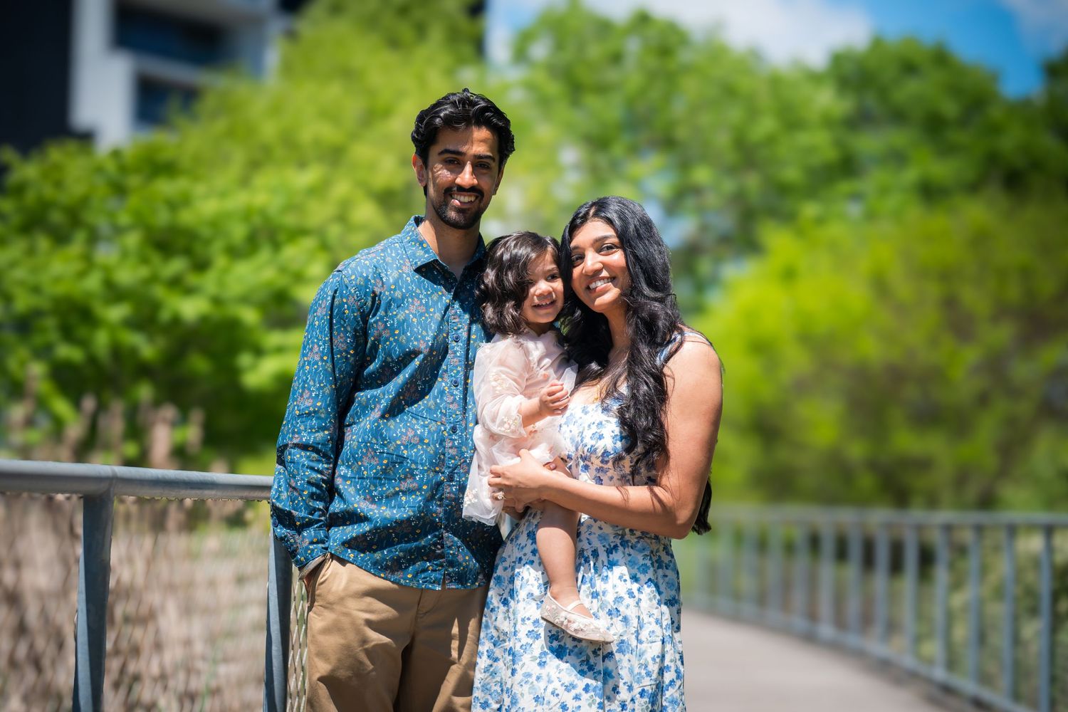 A family of three smiling on a bridge, with a man, woman, and young child in colorful spring outfits.
