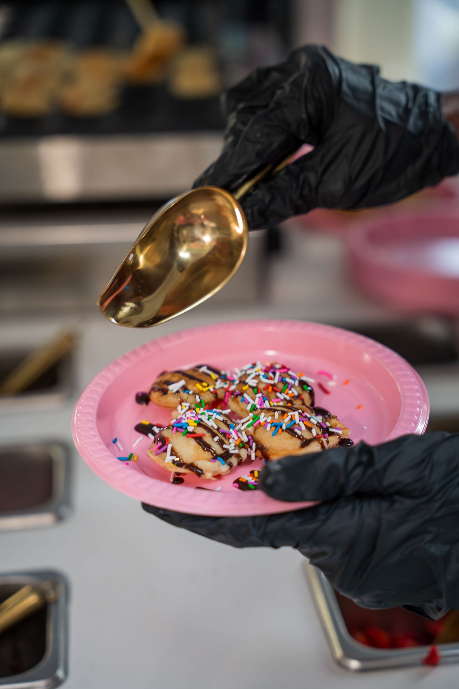 Dessert being garnished with colorful sprinkles on pink plates by gloved hands.