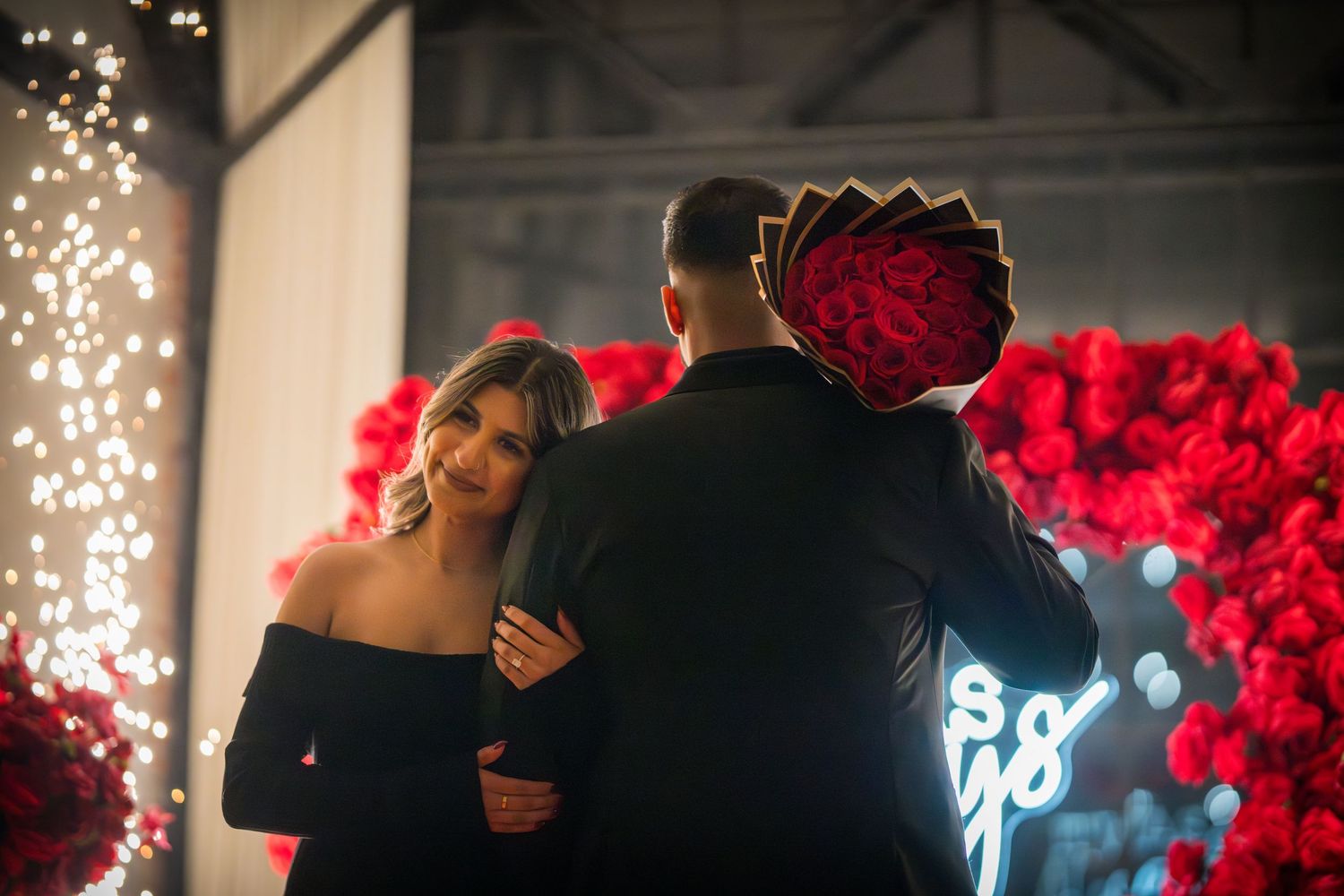 Silhouetted couple embracing against twinkling lights and red floral backdrop.