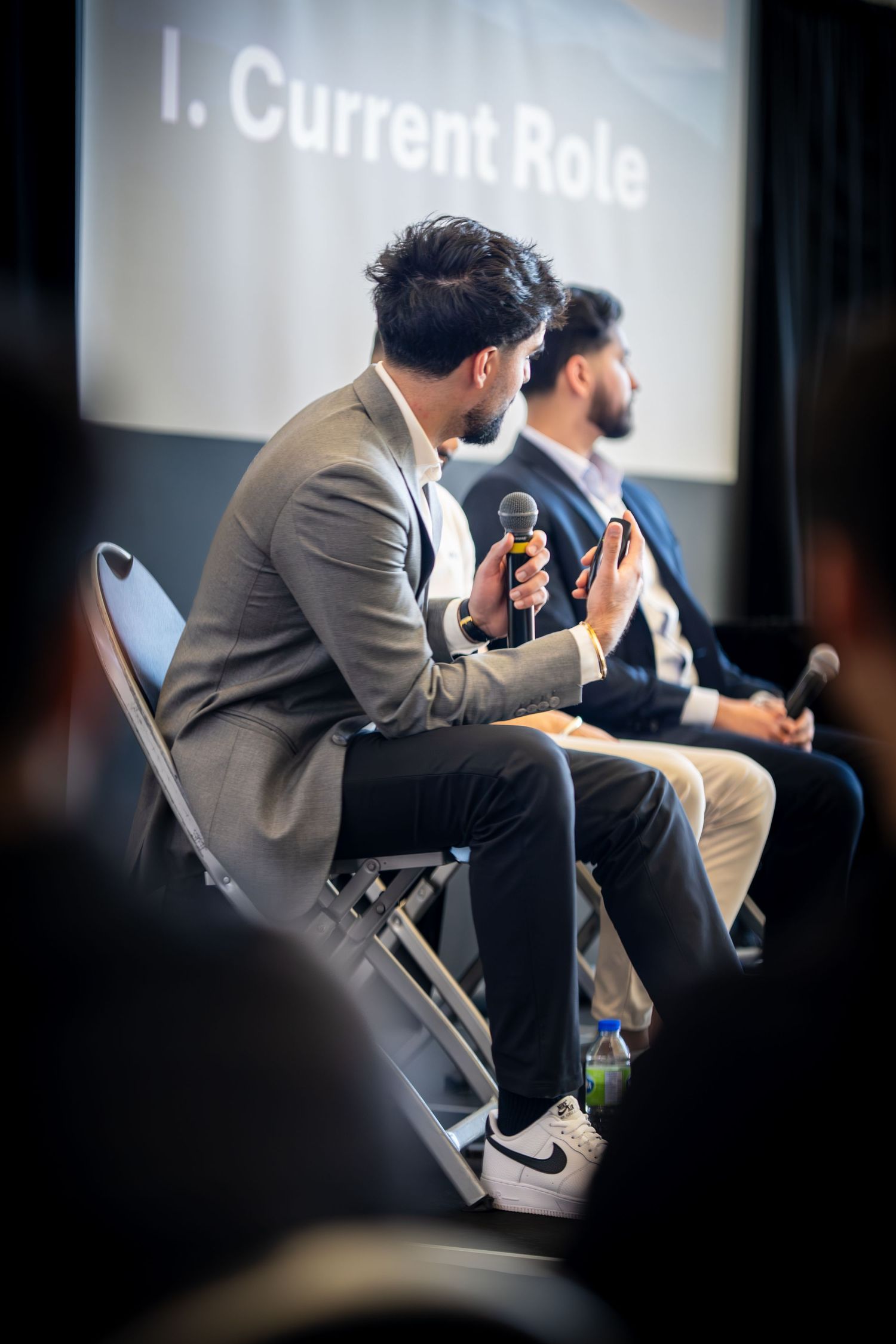 Speaker in grey blazer gestures while speaking into microphone during panel presentation.